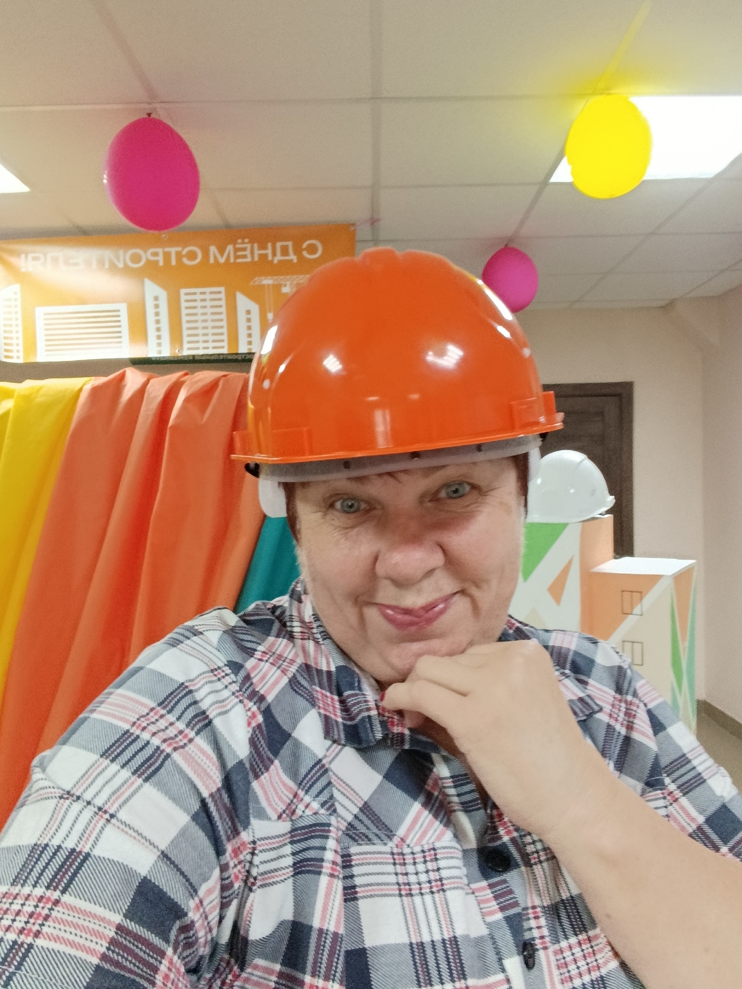 A woman wearing an orange hard hat and a plaid shirt is taking a selfie. She is indoors, in front of a backdrop of brightly colored fabrics in yellow, orange, and teal. In the background, a banner reads "С ДНЕМ СТРОИТЕЛЯ" (Happy Builder's Day), and two balloons, one pink and one yellow, are visible hanging from the ceiling. Another white hard hat is visible on a display stand to the right. The environment appears to be a room, possibly an office or event space, decorated for a celebration related to construction workers. The location is not specified as Svietlahorsk, Belarus.