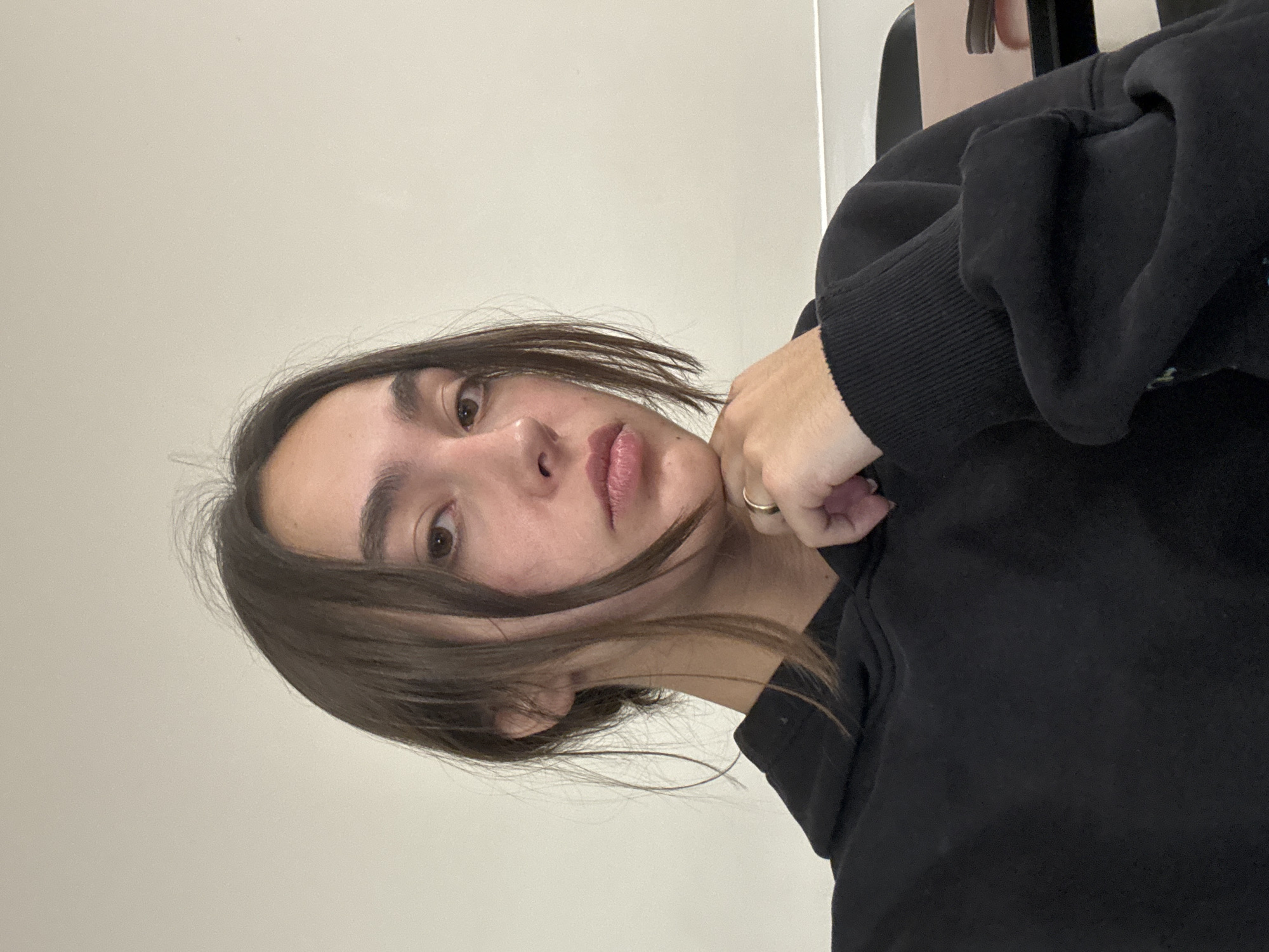 The image displays a close-up, selfie-style portrait of a young person, likely female, indoors. The subject has dark brown hair, casually styled and parted, with strands falling across her forehead and cheek. She looks directly at the camera with dark brown eyes and well-defined eyebrows. Her lips are full and have a natural, light pink tone, and her expression is neutral, possibly with a slight pout.
She is wearing a plain black long-sleeved top, appearing to be a hoodie or sweatshirt, with a visible ribbed cuff on her left sleeve. Her left hand is positioned near her chin or jawline, with fingers gently resting on her skin. A simple gold band ring is visible on one of her fingers.
The background consists of a plain, light-colored (off-white or beige) wall, indicating an indoor setting. A dark, indistinct object, potentially a piece of furniture like a chair back, is partially visible in the upper right corner of the frame. The lighting is soft and even, suggesting a well-lit interior without harsh shadows, likely during the daytime. The overall image is rotated 90 degrees clockwise, making the subject's head appear on the left side of the frame. There is no visible text in the image. The location context for the image is Huixquilucan de Degollado, Mexico.