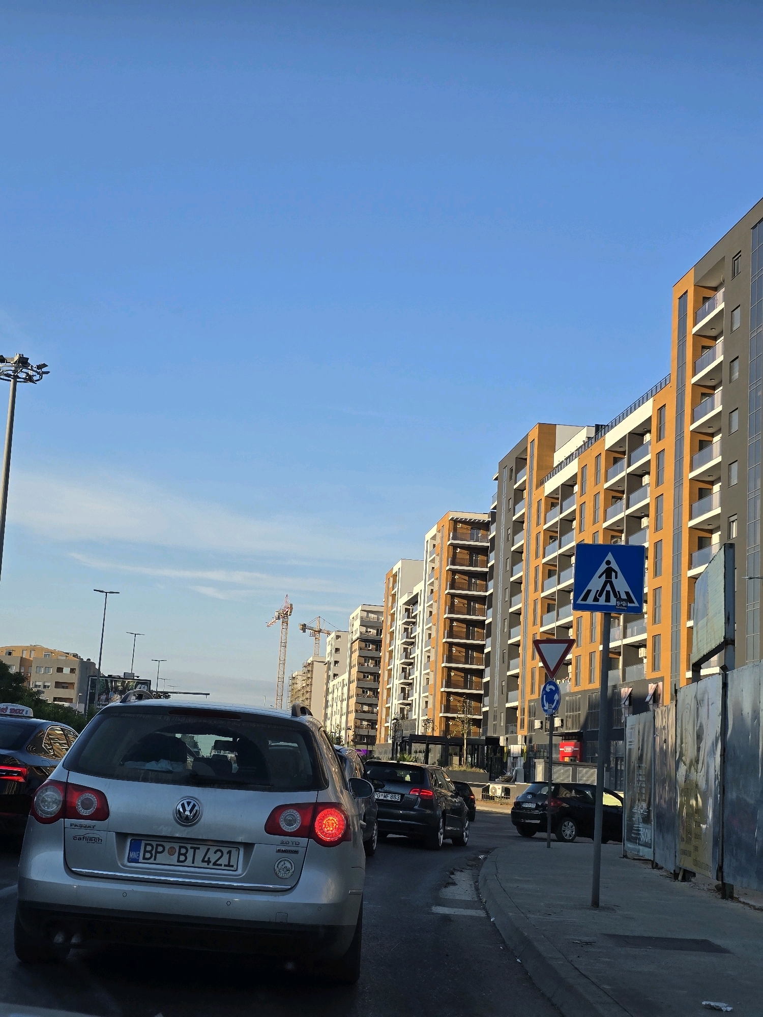 The image depicts a street scene in Podgorica, Montenegro, with a view looking down a street lined with modern, multi-story buildings. The sky is clear blue with a few wispy clouds. In the foreground, a silver Volkswagen Passat wagon is stopped in traffic, its license plate reading "MNE BP BT421". Other vehicles, including a black sedan and a smaller black car, are also visible in the traffic flow.
The buildings on the right side of the street are contemporary residential or office blocks, characterized by their orange and gray facades, numerous balconies with glass railings, and large windows. Construction cranes are visible in the background, suggesting ongoing development in the area.
On the sidewalk, a pedestrian crossing sign with a blue background and a white pictogram of a person walking is prominently displayed. Adjacent to it, a triangular yield sign and a circular roundabout sign are also present. The sidewalk appears to be made of concrete, and there is a metal fence or barrier on the right side of the image, which is covered in large posters or advertisements.
The lighting suggests it is daytime, likely late afternoon, given the quality of the sunlight casting shadows. The overall impression is one of urban development and daily life in a city.