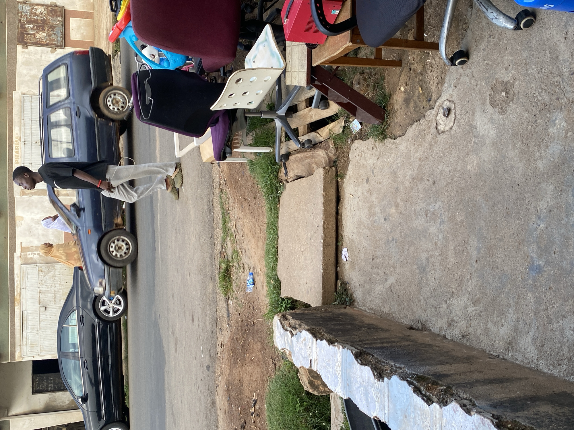 In Jos, Nigeria, on a street with a cracked, uneven concrete sidewalk, a young Black man in a black t-shirt and grey pants walks past a dark blue station wagon. Behind him, an older Black person in a tan hijab and a second person in a white hijab walk in the same direction, away from the viewer. The station wagon is parked on the street next to a black sedan. In the foreground, several office chairs are stacked on the sidewalk, including a purple and black chair, a white chair with a perforated back, and a red chair. A red plastic storage container sits on a wooden stool next to the chairs. The background shows a building with a beige facade and metal doors. The scene is illuminated by natural light, suggesting it is daytime.