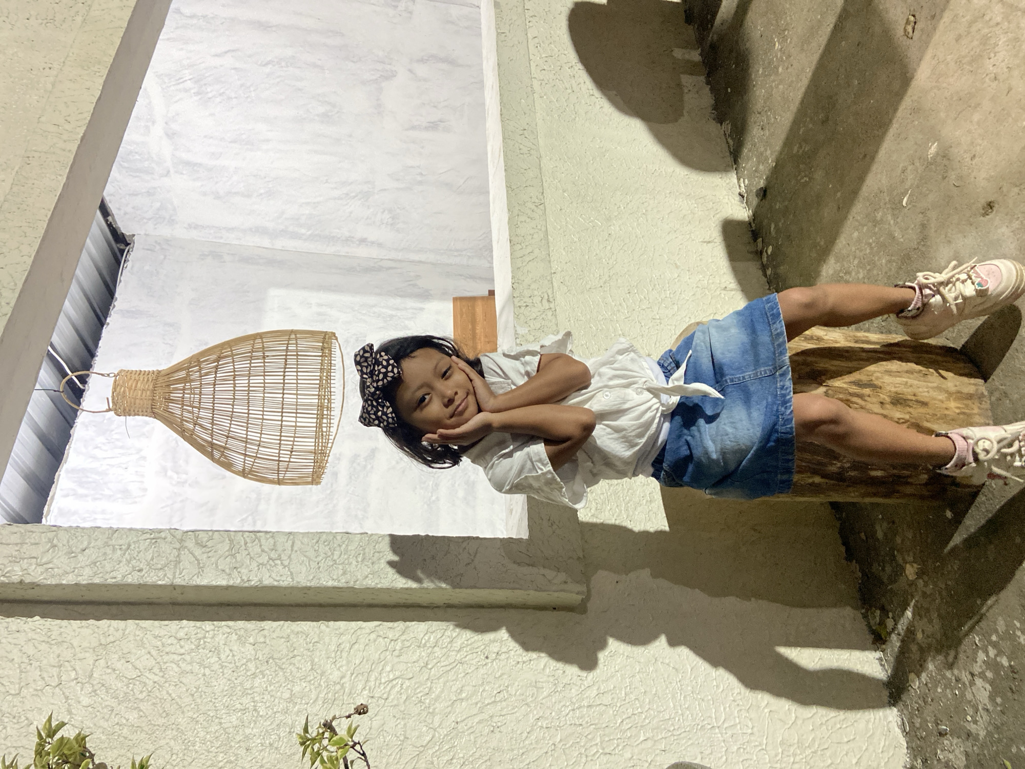 A young girl with dark hair and a black and white patterned bow is lying on her side on a log, propped up by her arms. She is wearing a white blouse and a denim skirt. She is looking towards the camera with a gentle smile and her hands are resting on her cheeks. The background consists of textured white walls, a woven light fixture, and a concrete floor. Shadows are cast on the floor, suggesting the presence of light from a source above and to the side, likely indicating daytime. The scene appears to be outdoors, possibly in a courtyard or a garden area. There is no visible text in the image. The overall mood is relaxed and peaceful.