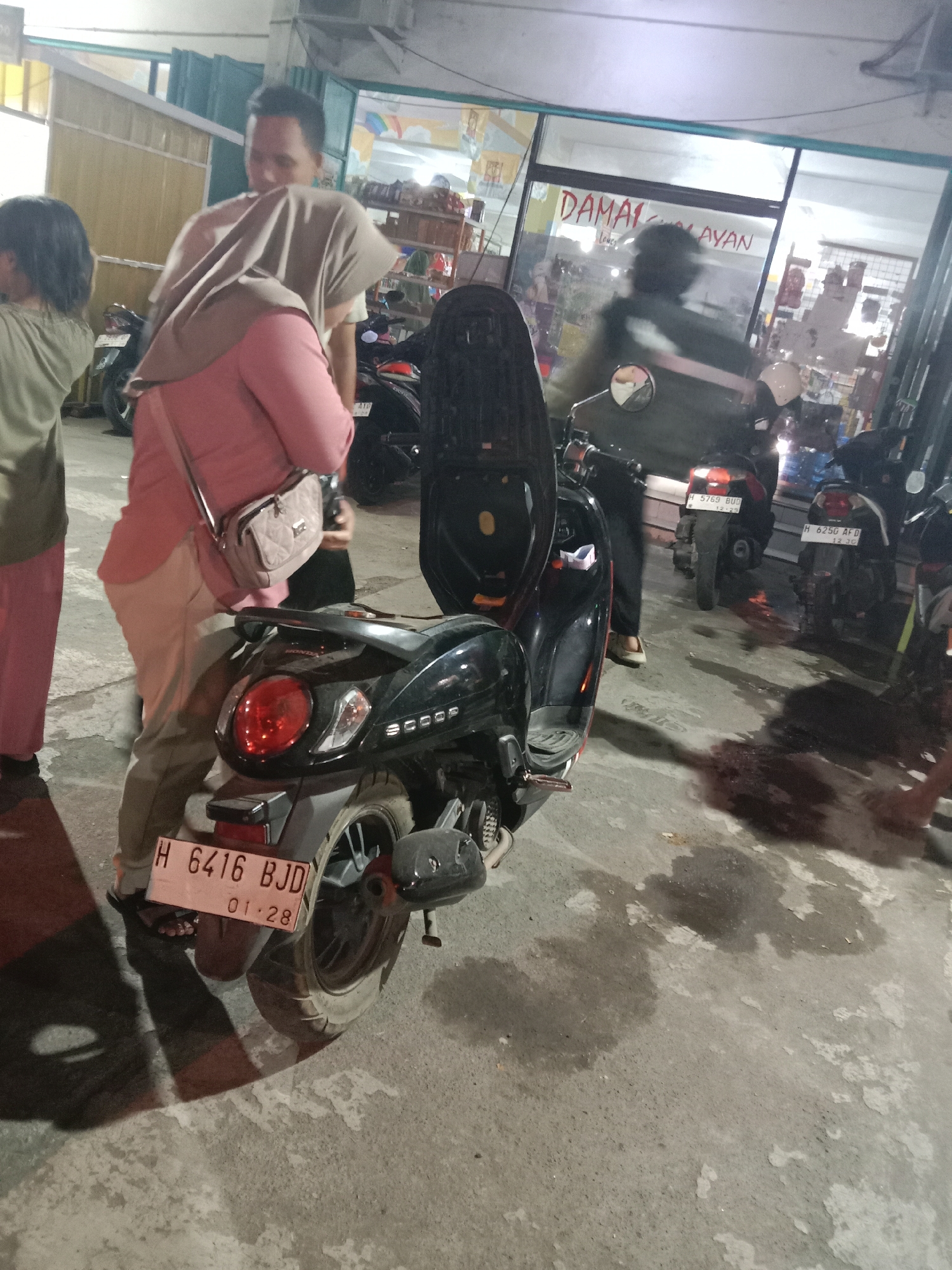 In Kebonsari, Indonesia, an outdoor scene depicts several motorcycles parked on a concrete surface at night. A dark-colored scooter with a red taillight is prominently featured in the foreground, its license plate reading "H 6416 BJD 01.28". A woman in a pink hijab and pink jacket, carrying a beige purse, stands near the scooter. A man in a light brown t-shirt is also present, looking towards the scooter. In the background, a shop window displays the words "DAMALAYAN" in red lettering. Other motorcycles are visible, including one with a license plate "H 5769 BUD 12.23" and another with "H 6250 AFD 12.30". The ground shows some wet patches, suggesting it might have rained or been recently washed. The lighting is dim, with artificial light illuminating the area.