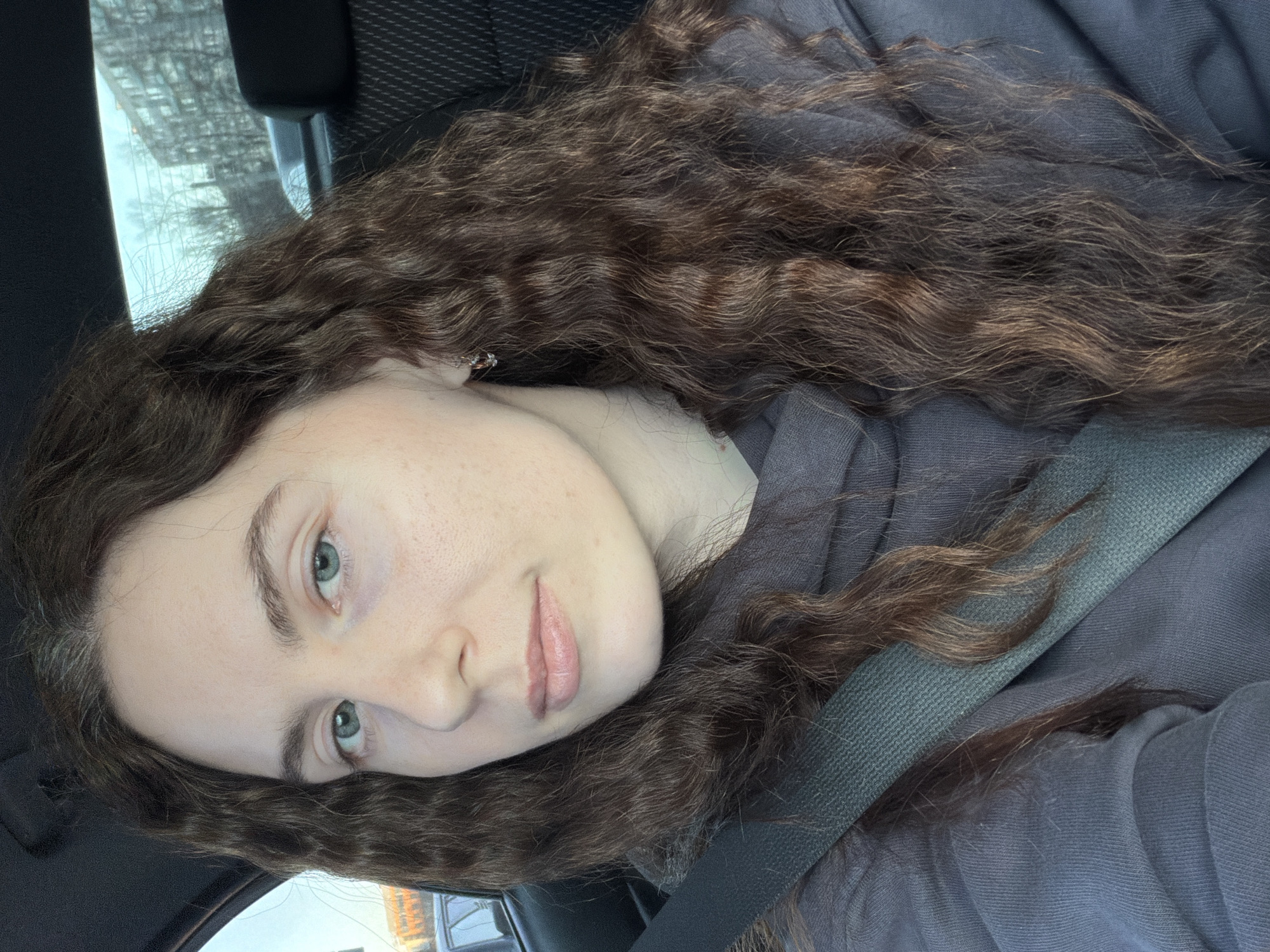 A woman with long, curly brown hair and light skin is in a car. She is wearing a grey sweatshirt and a seatbelt. The background shows a street scene with buildings. Given the visible architecture and foliage, it is likely a city in a northern latitude, but the exact location in Saint Petersburg, Russia, cannot be confirmed from the image alone.