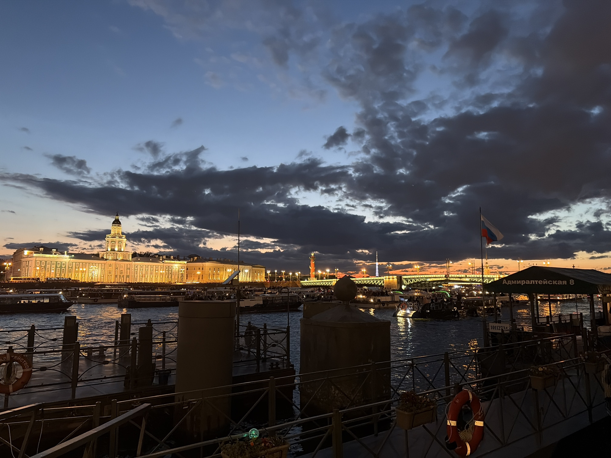The image depicts a waterfront scene in Saint Petersburg, Russia, during twilight. In the background, the Kunstkamera museum, illuminated by warm lights, stands prominently on the embankment of the Neva River. Further in the distance, the Rostral Columns and Palace Bridge are visible, also lit up. The sky is filled with dramatic, dark clouds against a gradient of blue and orange hues from the setting sun.
In the foreground, a pier or dock area is visible with metal railings and railings. Several boats, including tour boats and smaller vessels, are on the water, some with their lights on. Two large, cylindrical structures, possibly bollards or storage containers, are present on the dock. A red lifebuoy is attached to a railing. A green awning with the Russian text "Адмиралтейская 8" (Admiralteyskaya 8) is also visible, suggesting a pier location. The water reflects the lights of the city and the sky. A Russian flag is flying from a flagpole. People are visible on some of the boats and on the pier, observing the scenery. The overall atmosphere is one of a bustling evening by the river in a historic city.