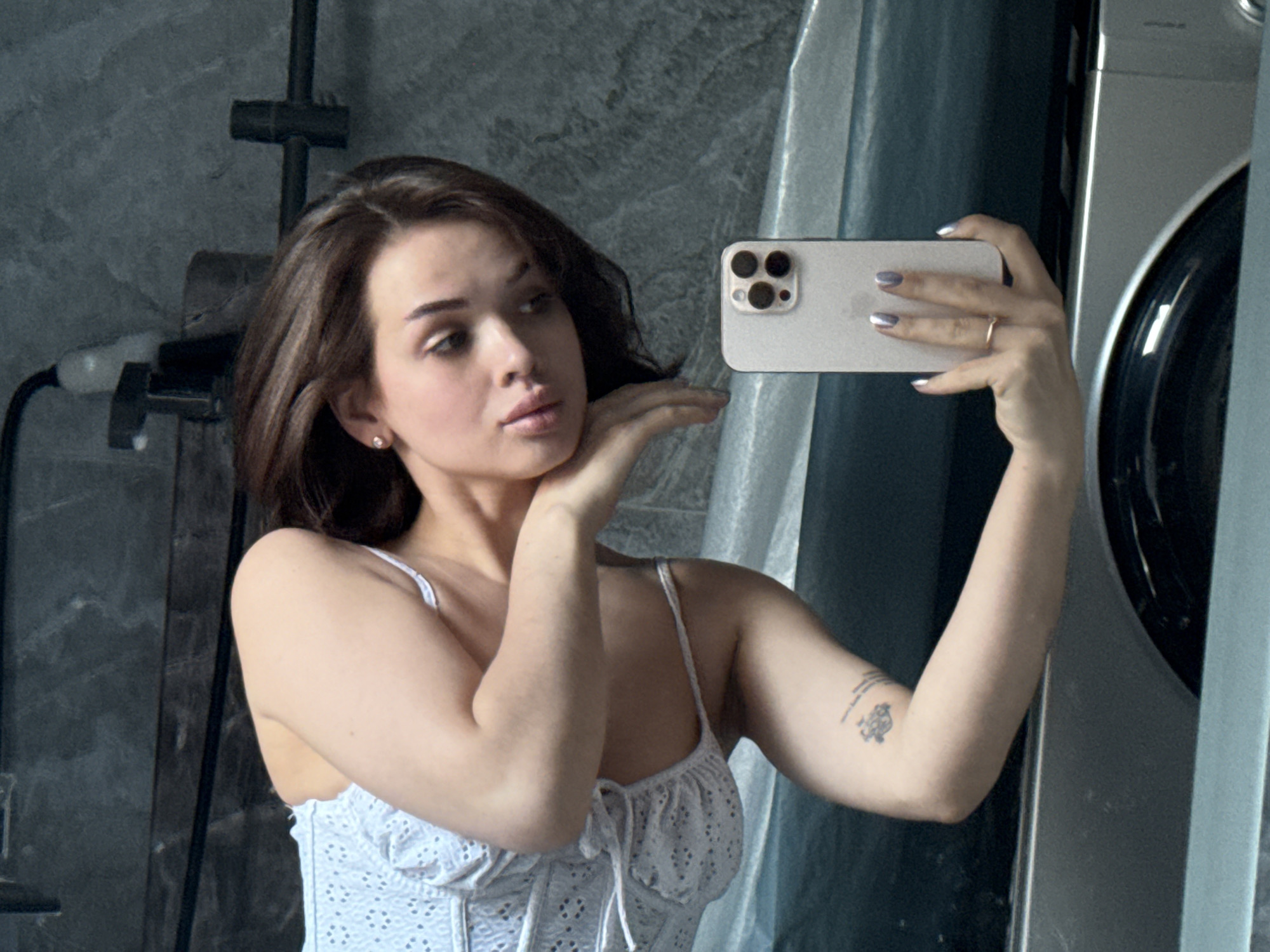 A fair-skinned woman with shoulder-length brown hair poses in a bathroom. She holds a white smartphone in her right hand, taking a selfie in front of a mirror, positioned in front of a washing machine. She is wearing a white top with delicate straps. Her left hand is positioned near her face. She has a ring on her ring finger and her fingernails are painted silver. A shower fixture is visible on the wall behind her. The image does not contain information to positively verify the location.
