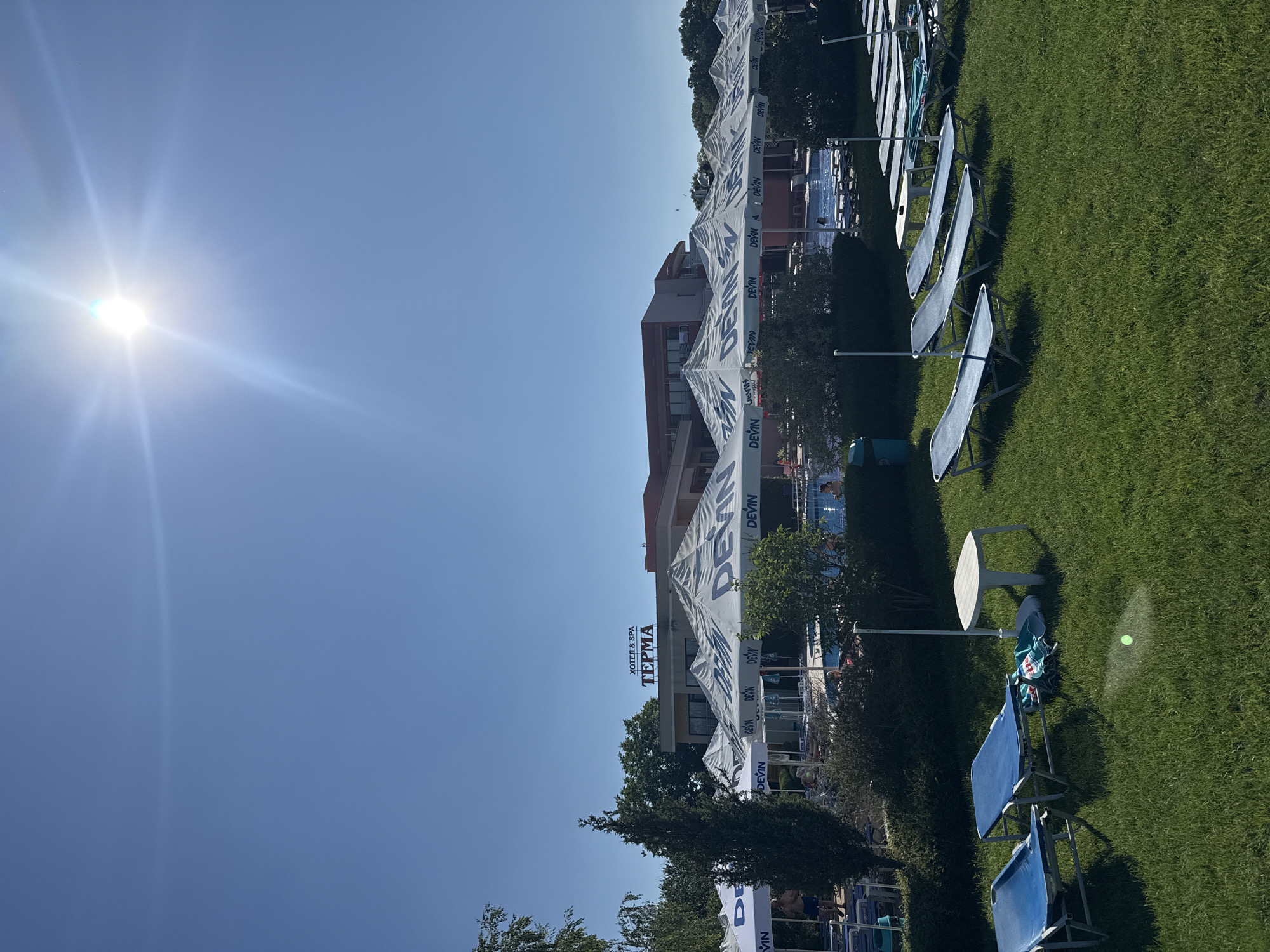 The image depicts an outdoor scene at the "HOTEL & SPA TERMA" in Yagoda, Bulgaria. In the foreground, green grass is mowed, with several blue lounge chairs arranged on it. A white plastic table stands between two of the lounge chairs. Behind the lounge chairs, a row of white umbrellas with blue lettering that reads "DEVIN" are set up. These umbrellas provide shade over more lounge chairs. In the middle ground, a swimming pool is visible, with people swimming and relaxing. Behind the pool area and umbrellas, the hotel building is visible, with "HOTEL & SPA TERMA" signage displayed prominently on its facade. The sky is clear and blue, with the sun shining brightly and creating lens flare. Trees and bushes surround the hotel and pool area, indicating a resort or hotel setting.