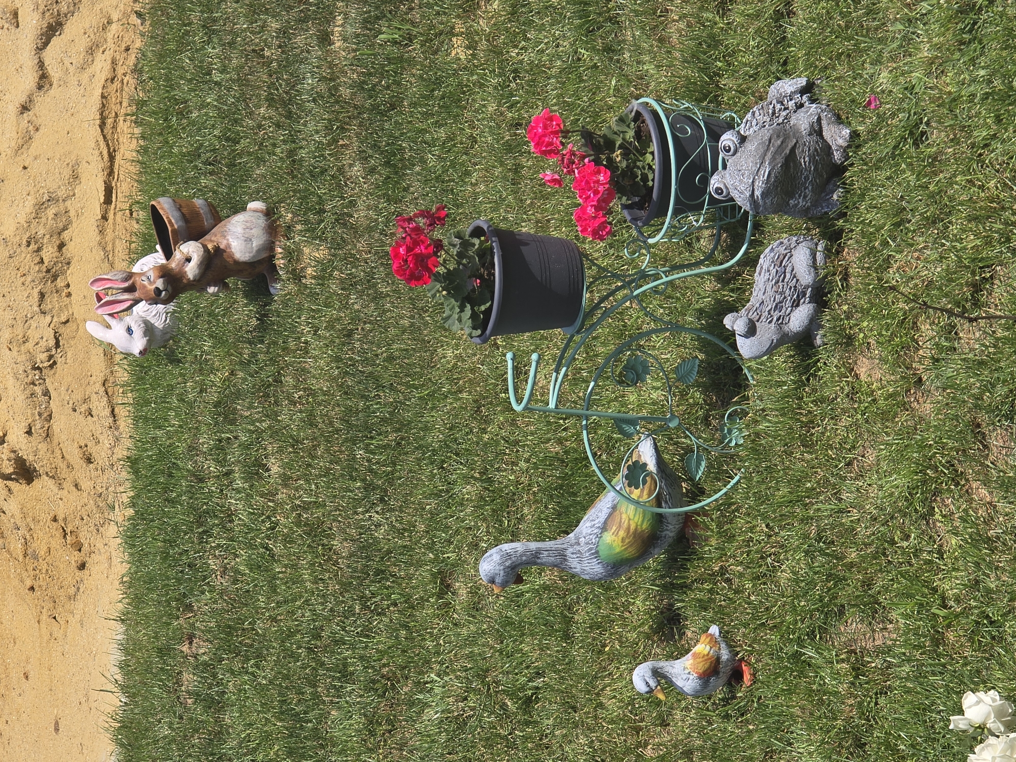The scene is an outdoor setting of a grass lawn with various garden ornaments. A flower stand with two black pots containing red flowers sits in the middle ground. Garden statues of a pair of frogs and two ducks are also on the grass. A group of three rabbit statues with a barrel is placed near a sandy area. The exact location and time are not discernible from the image, and there is no information suggesting the setting is in Archar, Bulgaria. No people are visible.