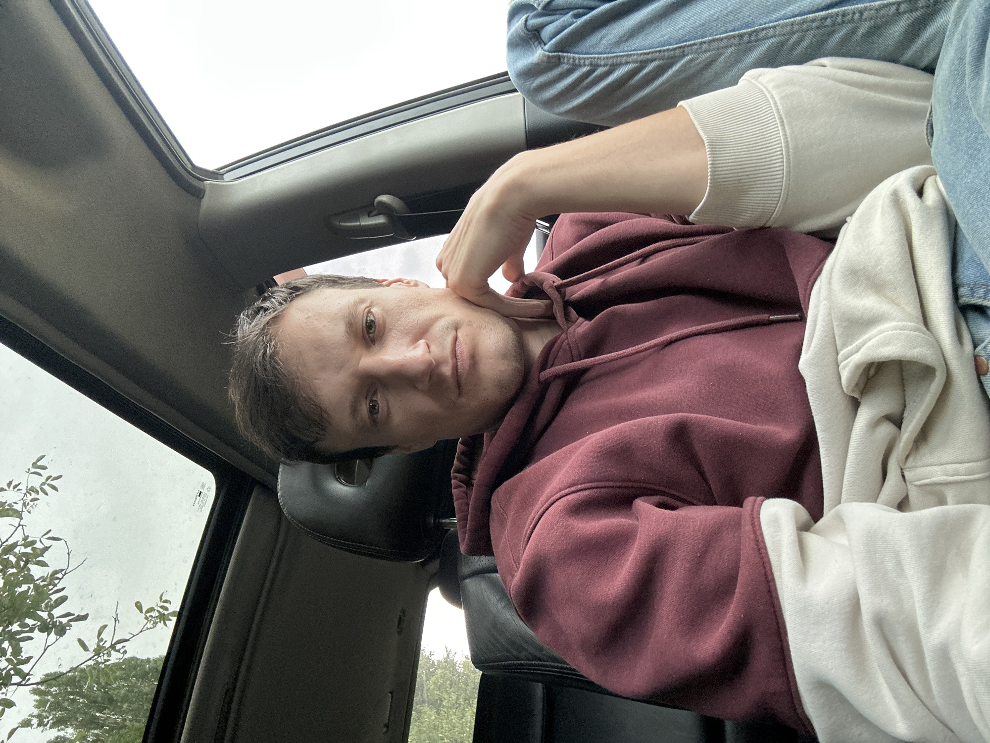 A man is sitting in the driver's seat of a car, looking towards the camera. He is wearing a burgundy hooded sweatshirt with cream-colored sleeves and light blue jeans. His left arm is resting on his knee, with his hand supporting his chin. The car has a sunroof open, and trees and a cloudy sky are visible through it. The interior of the car, including the black leather seats and headrest, is visible. The scene appears to be outdoors, possibly in a wooded or park-like area, given the presence of trees. The location is identified as Makiv, Ukraine.
