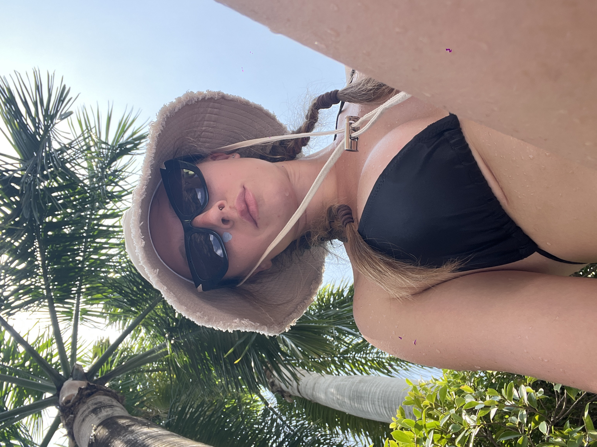 The image shows a young woman from a low-angle perspective, wearing a black bikini, a light-colored bucket hat, and sunglasses. She has her hair in two braids and is looking up towards the camera. Palm trees and lush green foliage are visible in the background, suggesting an outdoor, tropical environment, likely a resort or a pool area. The woman's skin appears wet, indicating she may have just been swimming. The bright sky suggests it is daytime. The scene is consistent with a location in Bang Sao Thong Subdistrict, Thailand, which is known for its tropical climate and popular tourist destinations with such natural scenery.