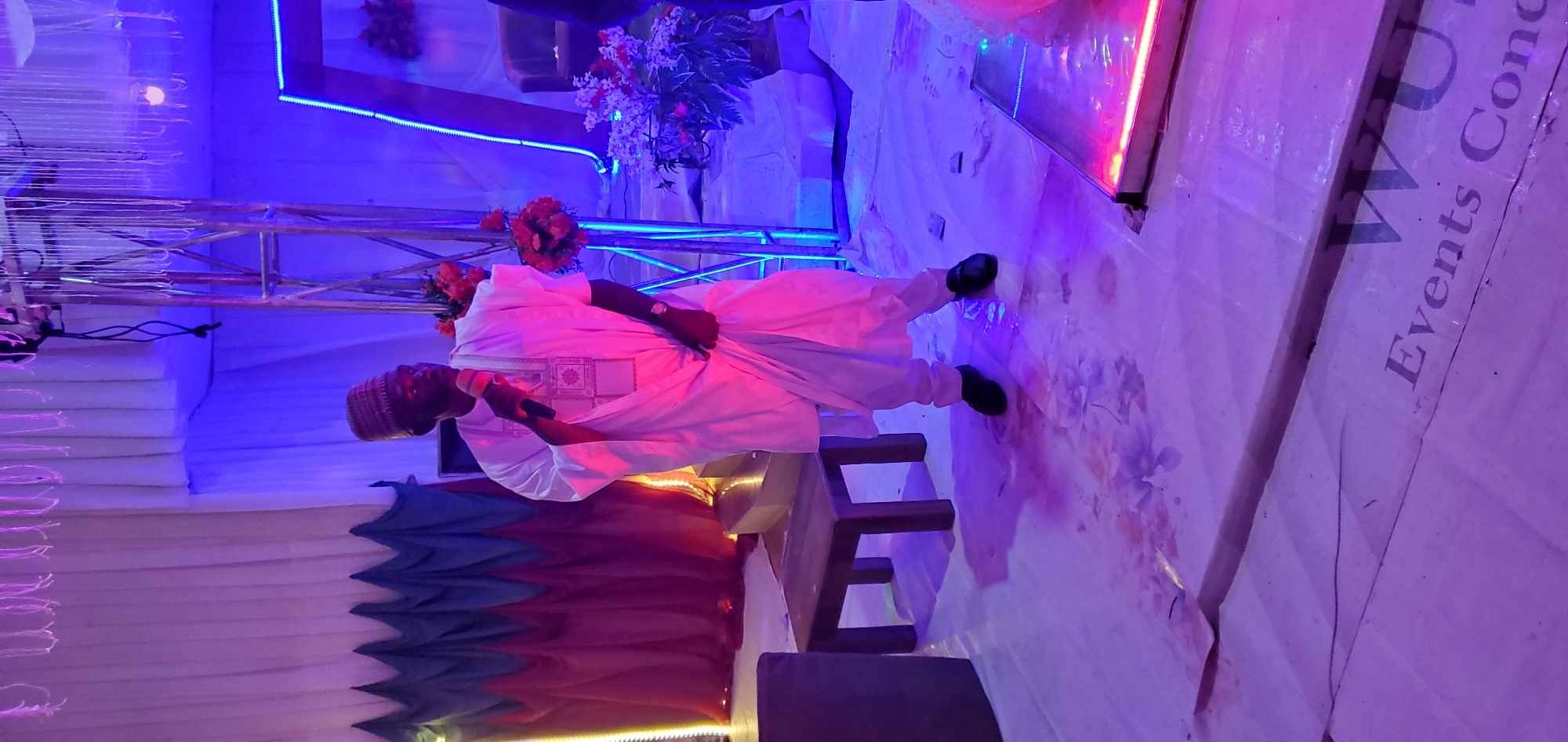 This image depicts an indoor event space, configured for a presentation or performance. A fair-skinned adult male of apparent African descent stands centrally, addressing an audience while holding a red and black microphone to his mouth. He is dressed in traditional light-colored attire, including an embroidered robe, a woven cap, and black dress shoes, with a watch on his left wrist.
The floor is covered with a white, sheet-like material featuring subtle floral patterns and noticeable dark pinkish-red stains, particularly under and around the man's feet. Visible seams and creases traverse the floor covering. To the man's left, a small dark wooden stool or table is present, along with a partial view of a dark cushioned seat.
The background is adorned with event decorations and structures. On the left, a draped backdrop in white, blue, and red/maroon hues is arranged in a scalloped pattern. To the right, a metal truss structure is decorated with red and orange floral arrangements. Further right, a cluster of white and pink flowers is visible next to a rectangular frame outlined with a blue neon-like light strip. Numerous thin, white string decorations descend from above, and a low-profile, illuminated rectangular platform rests on the floor at the far right. The scene is bathed in theatrical lighting, casting strong pink/red and blue hues across the space. The specific city of Lankan, Nigeria cannot be visually confirmed from the details within the image.
