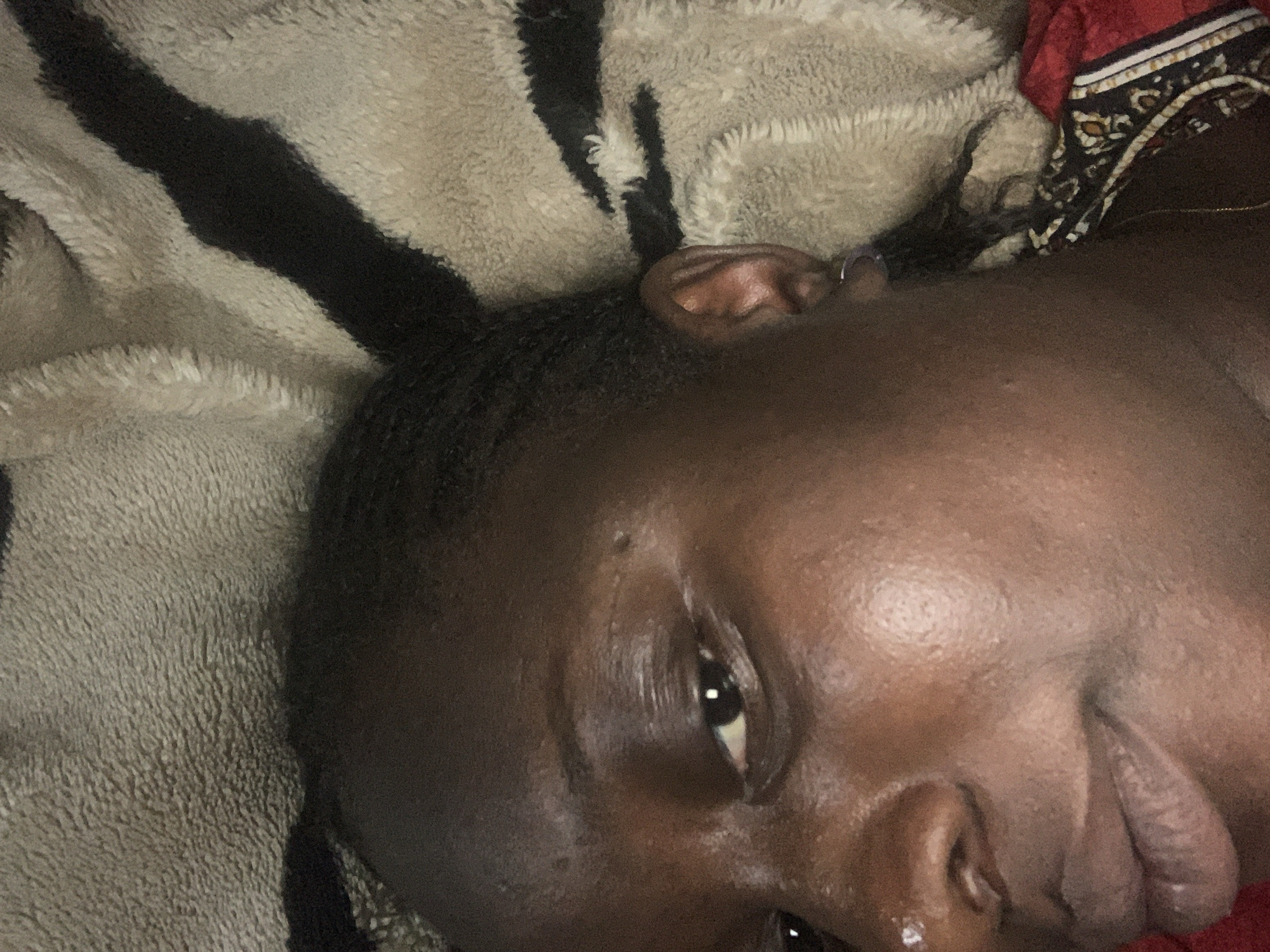 A close-up shot shows the upper portion of a person's face, from the forehead to the chin, as they lie on a blanket with a black and white striped pattern. The person has dark skin and braided hair. They are wearing a small, dark earring in their visible ear. The lighting is dim, creating a soft focus on the face. The blanket appears to be made of a plush, textured material. The background is out of focus and is dominated by the pattern of the blanket.