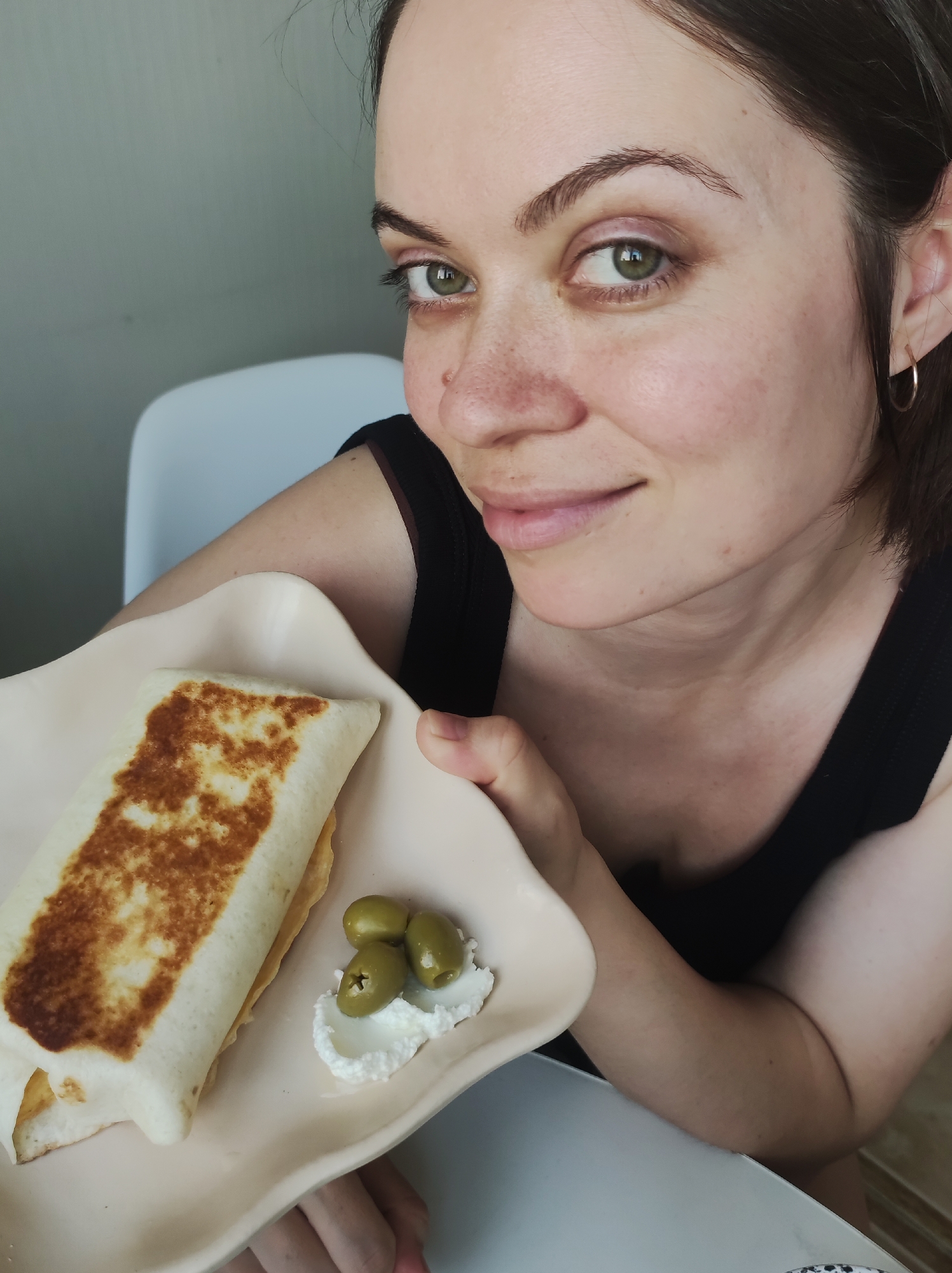 A woman is shown from the chest up, holding a beige plate with food. The plate contains a rectangular burrito with grill marks, three green olives, and a small dollop of white cream. The woman has fair skin, freckles, and dark hair pulled back from her face. She is wearing a black tank top and a small gold hoop earring. The background includes a cream-colored wall, a white chair, and the edge of a table. The location is unknown.