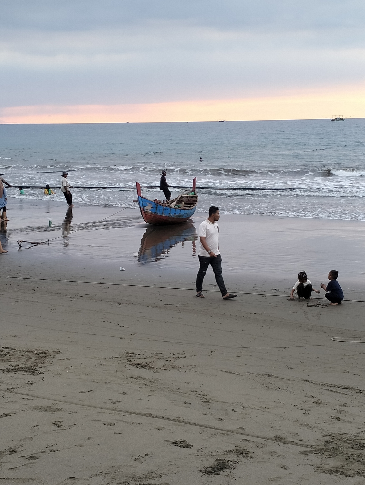 The image depicts a beach scene in Uleekareung, Indonesia, with the ocean in the background and a sandy shore in the foreground. The sky is overcast with hues of gray and faint pink from the setting sun. Several people are present on the beach.
In the middle ground, a blue and red wooden boat is pulled ashore by several individuals. Two men, one in a white shirt and dark pants, the other in a dark shirt and pants, are actively pulling on ropes attached to the boat. A third person in a dark shirt is also near the boat, potentially assisting. Two young children are playing in the sand near the water's edge, closer to the foreground. A man in a white shirt and dark pants is walking across the sand, looking towards the children. Further in the background, several other figures are visible near the water, and two fishing boats are visible on the horizon. The wet sand reflects the sky and the boat, indicating the tide is out or receding.