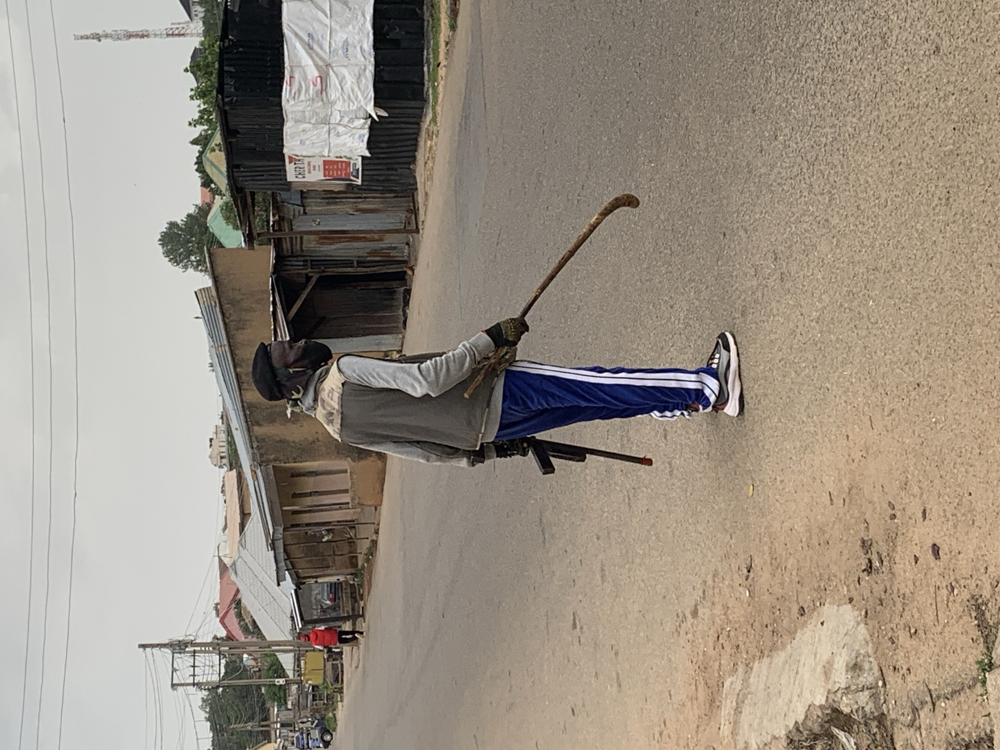 A person is standing on a paved road in what appears to be Jos, Nigeria. They are wearing a gray jacket, blue sweatpants with white stripes, and athletic shoes. The person is holding a long, curved wooden stick in their right hand and what appears to be a rifle in their left hand. Buildings line the road, including structures made of corrugated metal and concrete. A communications tower is visible in the background. Other people and vehicles are seen further down the road. The sky is overcast.
