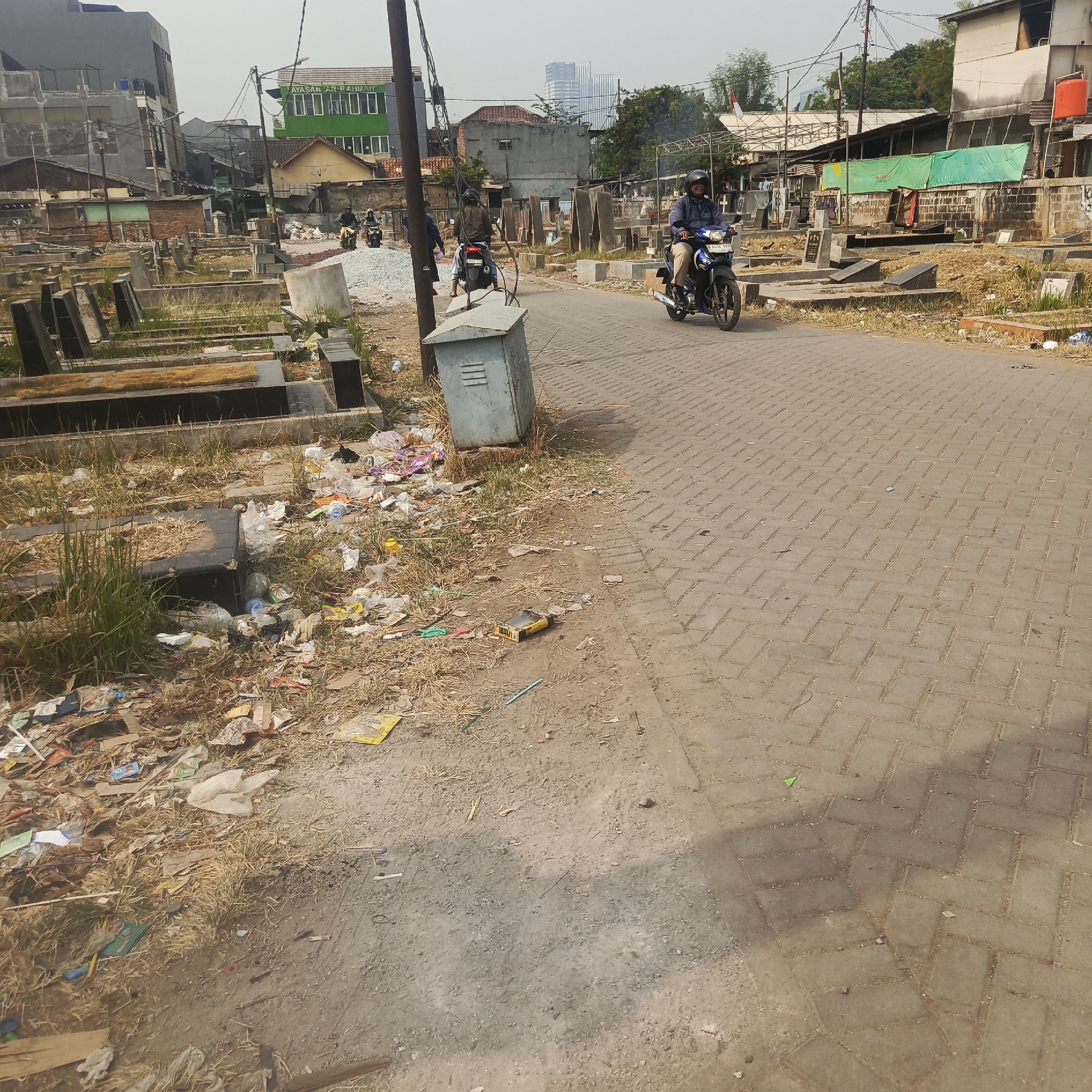 In South Jakarta, Indonesia, a paved road runs through a cemetery dotted with gravestones. The road is made of interlocking concrete pavers. On the left side of the road, the cemetery is visible, with many gravestones and dry grass. Scattered trash is present in the cemetery, including plastic bags, bottles, and paper. On the right side of the road, a person on a blue motorcycle is riding towards the camera. They are wearing a helmet and a blue jacket. Further down the road, other motorcycles and people are visible. In the background, buildings of various sizes and conditions are seen, along with trees and utility poles. The sky is overcast.