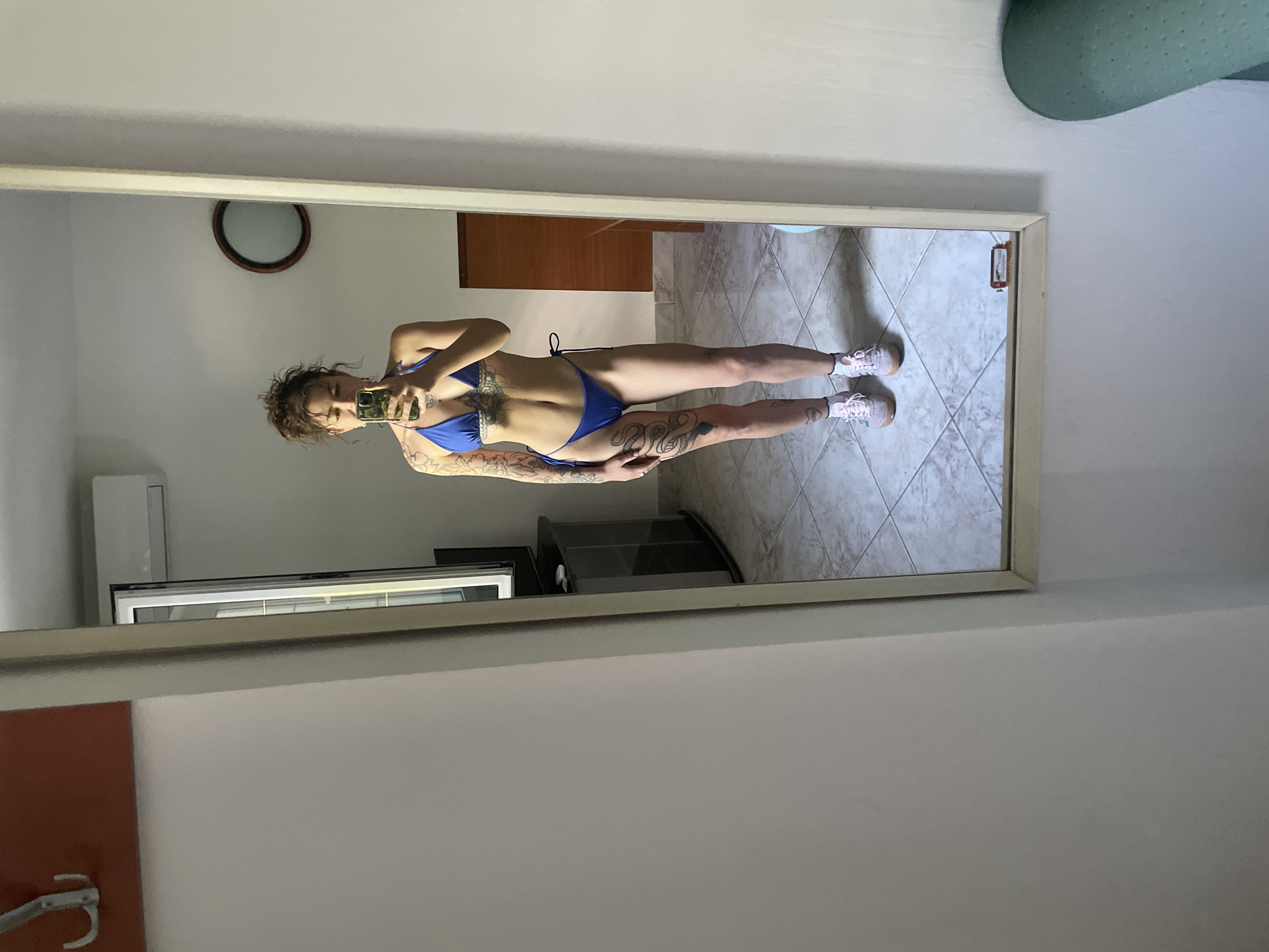 A full-length, front-facing mirror reflects a woman standing indoors, taking a selfie with her phone. She is wearing a blue bikini and pink sneakers. Her body is covered in various tattoos, including a snake design on her left leg. The room appears to be a bedroom or living space, with tiled flooring, a wooden door in the background, and an air conditioning unit mounted on the wall. A circular mirror with a dark frame is visible on the wall behind her. The scene is well-lit, suggesting daytime. The location is identified as Herceg Novi, Montenegro.