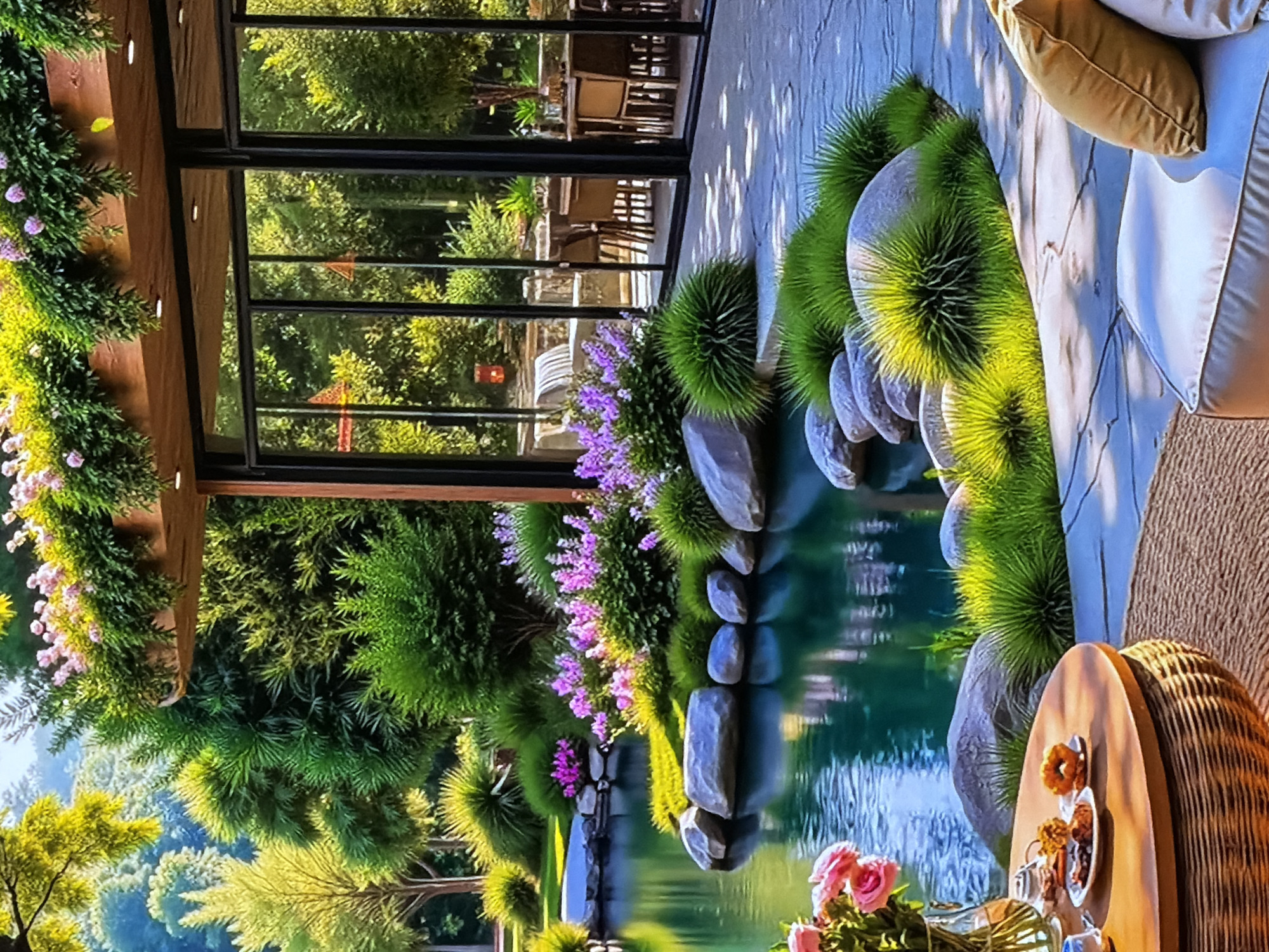 This image captures a serene outdoor patio or veranda seamlessly integrated with a lush garden, bathed in bright daylight. The scene, located in Sofiivska Borshchahivka, Ukraine, suggests a tranquil setting for relaxation or dining.
In the foreground, a comfortable outdoor sofa with light-colored cushions and a mustard-yellow throw pillow invites repose. Adjacent to it, a small round wooden table with a woven base holds two white plates of donuts or pastries, a glass vase with fresh pink roses, and other refreshments, implying an inviting leisure or dining activity.
The patio extends towards a beautifully landscaped garden featuring a tranquil body of water, possibly a pond or stream. Its banks are meticulously designed with smooth grey rocks and distinctive tufts of vibrant green, spiky ornamental grass. Clusters of purple flowering plants add splashes of color along the water's edge.
To the left, a wooden overhead structure or pergola is adorned with cascading green foliage and delicate pink flowers, providing a natural canopy. A modern glass wall with dark frames separates the outdoor area from an interior space, through which the reflections of the surrounding greenery and partial views of tables and chairs, possibly belonging to a cafe or restaurant, are visible.
Beyond the immediate garden, a dense expanse of various trees and foliage creates a rich, verdant backdrop. The overall impression is one of peaceful natural beauty and refined outdoor living. The time of day appears to be sunny, with clear weather. No people or visible text are present in the image.