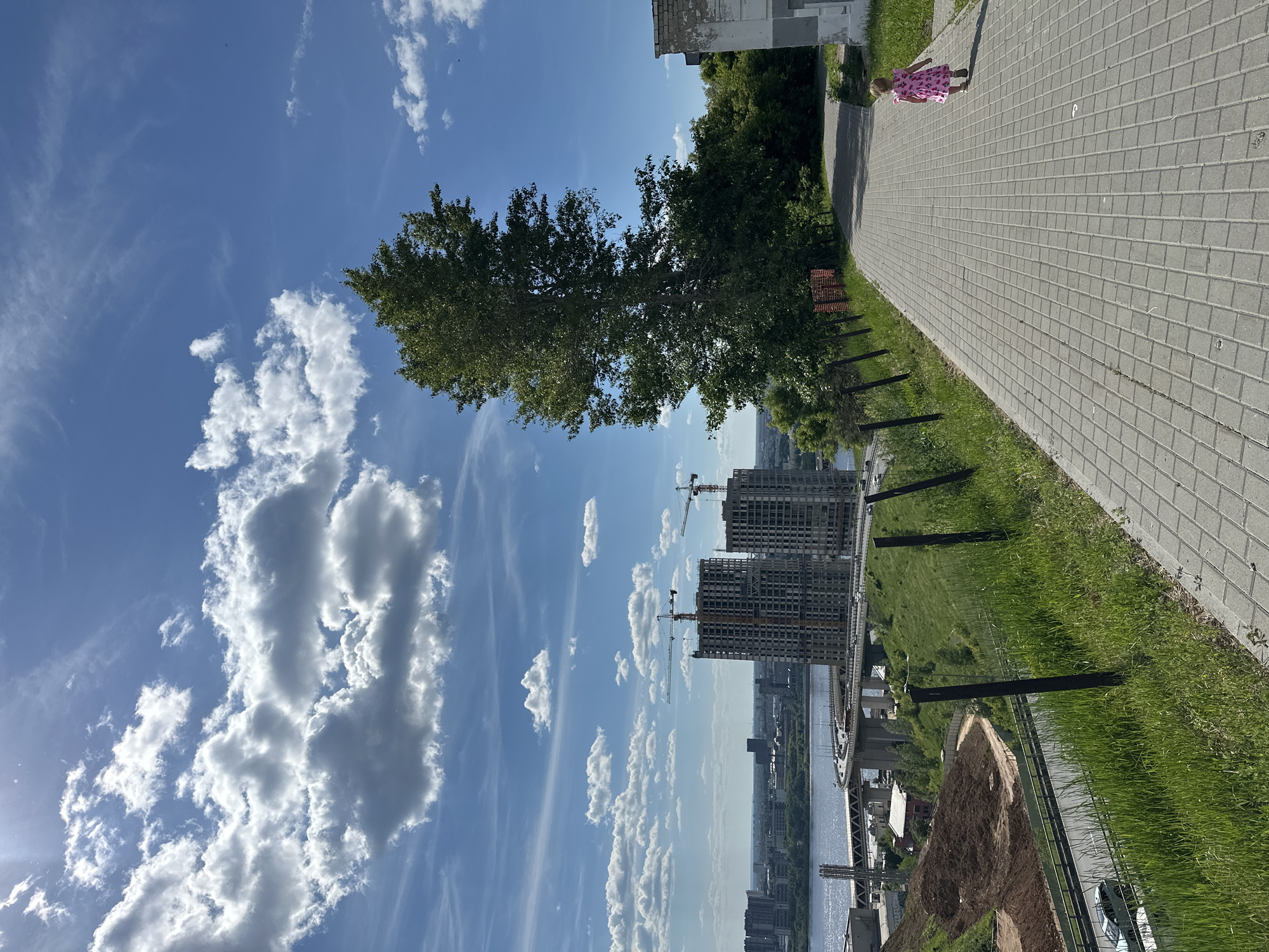 This image captures a wide-angle outdoor scene from Fedorovsky Embankment in Nizhny Novgorod, Russia, looking northwest towards the confluence of the Oka and Volga rivers. The weather is sunny with a bright blue sky and scattered white cumulus clouds.
In the immediate foreground on the right, a paved pedestrian walkway made of grey square tiles extends diagonally from the bottom right towards the mid-left. A small child, wearing a pink floral dress, walks away from the camera on this path. To the right of the path, a partially visible, dilapidated light-colored building with peeling plaster and visible brickwork is present. On the left foreground, a steep grassy slope with lush green vegetation descends sharply. Dark, weathered wooden fence posts are spaced along the edge of this slope, bordering the walkway.
In the midground, a tall, mature deciduous tree with dense green foliage stands prominently to the left of the paved path. Below the grassy slope, a multi-lane road is visible, with at least one light-colored car driving on it. Further down the slope, an area of exposed brown earth, consistent with excavation or a construction site, is visible. Beyond this, an elevated, multi-span concrete bridge structure, part of the Kanavinsky Bridge, curves across the scene, carrying vehicular traffic over the Oka River.
The background features the wide expanse of the Oka River, reflecting the sunlight and appearing light blue-grey. On the far bank of the river, two tall, unfinished residential buildings, part of the "Volzhskiye Zori" complex, are under construction. Each building has multiple completed concrete stories and is topped with bright yellow tower cranes. Beyond these construction sites, the urban skyline of Nizhny Novgorod extends, featuring various buildings of different heights and styles, stretching towards the horizon.