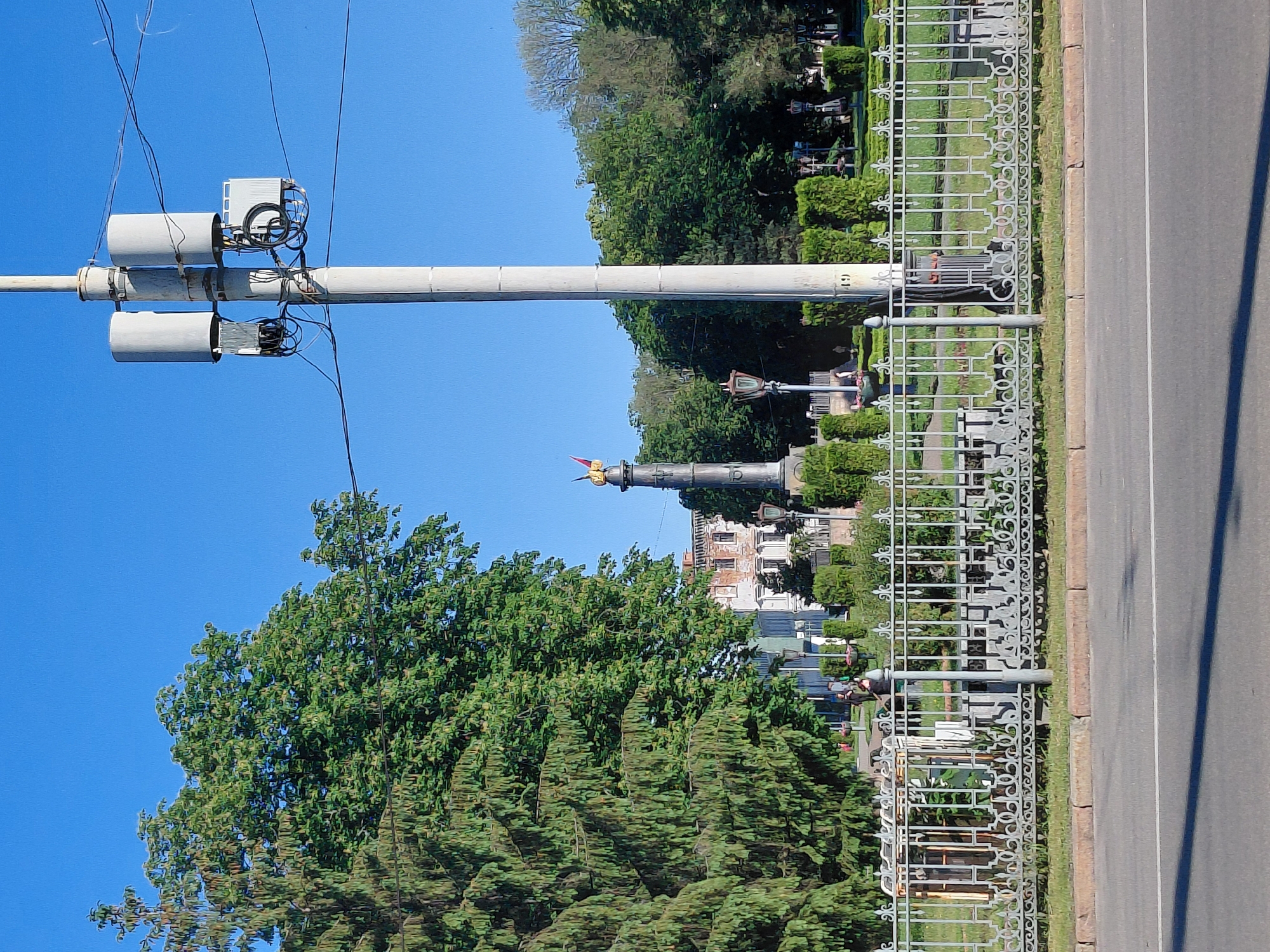An outdoor scene captured on a clear, sunny day depicts a street bordering a manicured park or garden. The bright blue sky dominates the upper portion of the image, suggesting mid-morning or afternoon.
In the foreground, a prominent tall, light-colored utility pole stands vertically on the left. Attached to this pole are several electronic devices: two grey cylindrical units and two rectangular units, all connected by a web of black wires. These devices are mounted on horizontal arms extending from the main pole. Numerous thinner black wires crisscross the sky from these installations. On the main pole, near its base, the number "49" is visible vertically, and on one of the horizontal arms, the letters "G1" are discernible.
To the right of the pole is an asphalt road with a single white dividing line. A concrete curb separates the road from a strip of grass. A distinct shadow, likely cast by the pole, stretches across the road.
Separating the road from the park is an ornate grey metal fence featuring intricate scrollwork and grid patterns. Beyond this fence, the park displays lush green foliage, including well-maintained hedges and various trees, both leafy deciduous and coniferous.
In the mid-ground, nestled among the trees, a tall, dark-colored monument resembling an obelisk or a column is visible. It is topped with a golden, possibly winged, figure. A traditional-style lamppost with a dark lantern fixture is also visible within the park area, near the monument. In the distant background, beyond the park and monument, parts of multi-story buildings with visible windows can be seen.
No people or specific activities are clearly visible in the image, suggesting a tranquil and static urban landscape.