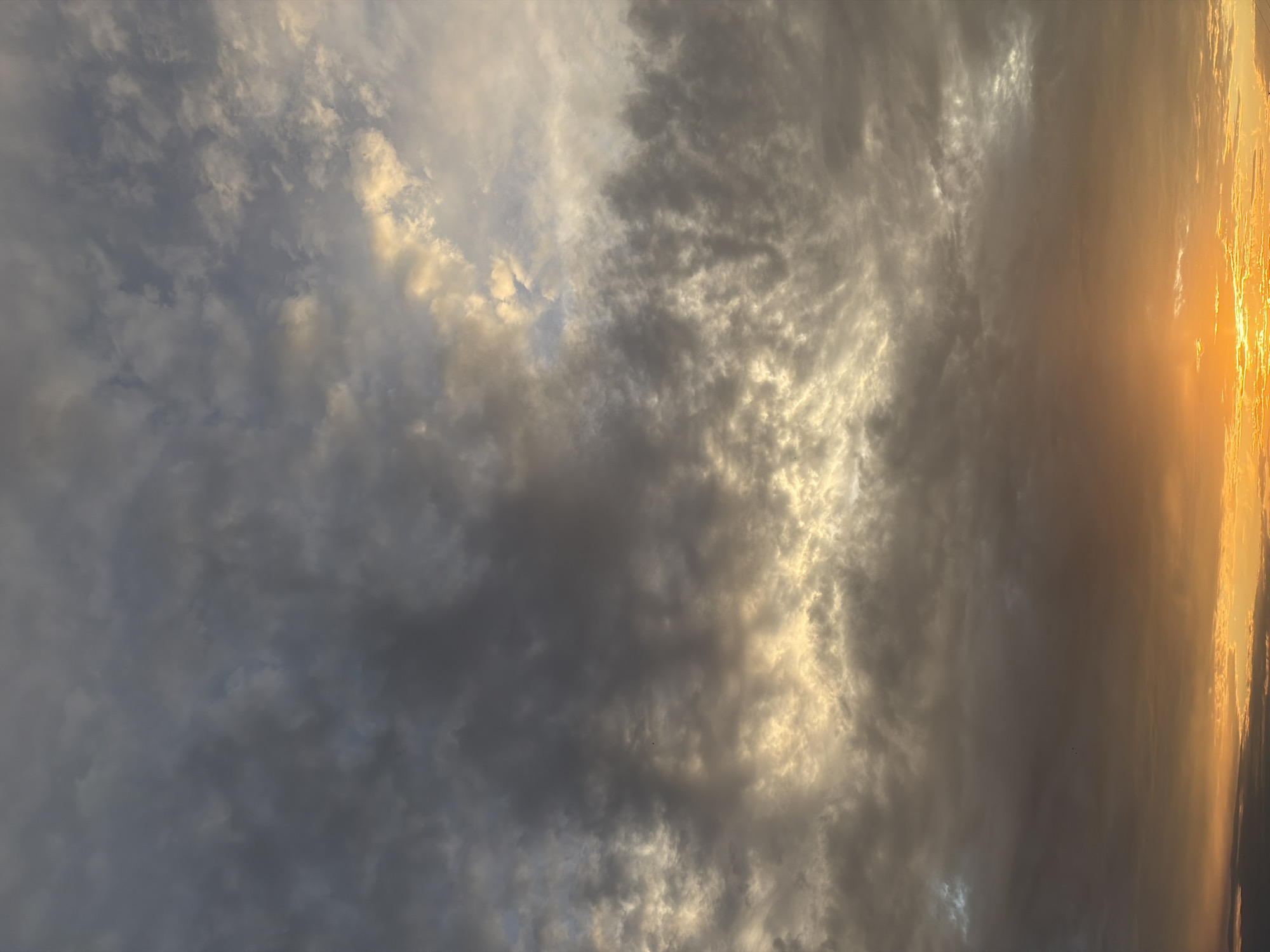 A photograph captures a sky filled with a gradient of colors from a vibrant sunset to dark, brooding clouds. The bottom of the image is dominated by a bright orange and yellow glow of the setting sun, which is partially obscured by thin, horizontal clouds. Above this, the sky transitions into a mix of grays and whites as thicker clouds dominate the mid-section, with some areas illuminated by the fading sunlight, creating a luminous effect. The upper portion of the sky is a darker, more muted gray, indicating the approaching twilight. No specific landmarks or elements of the city of Chaikovsky, Russia are visible in this purely atmospheric composition. The image appears to be taken from a ground-level perspective, looking upwards.