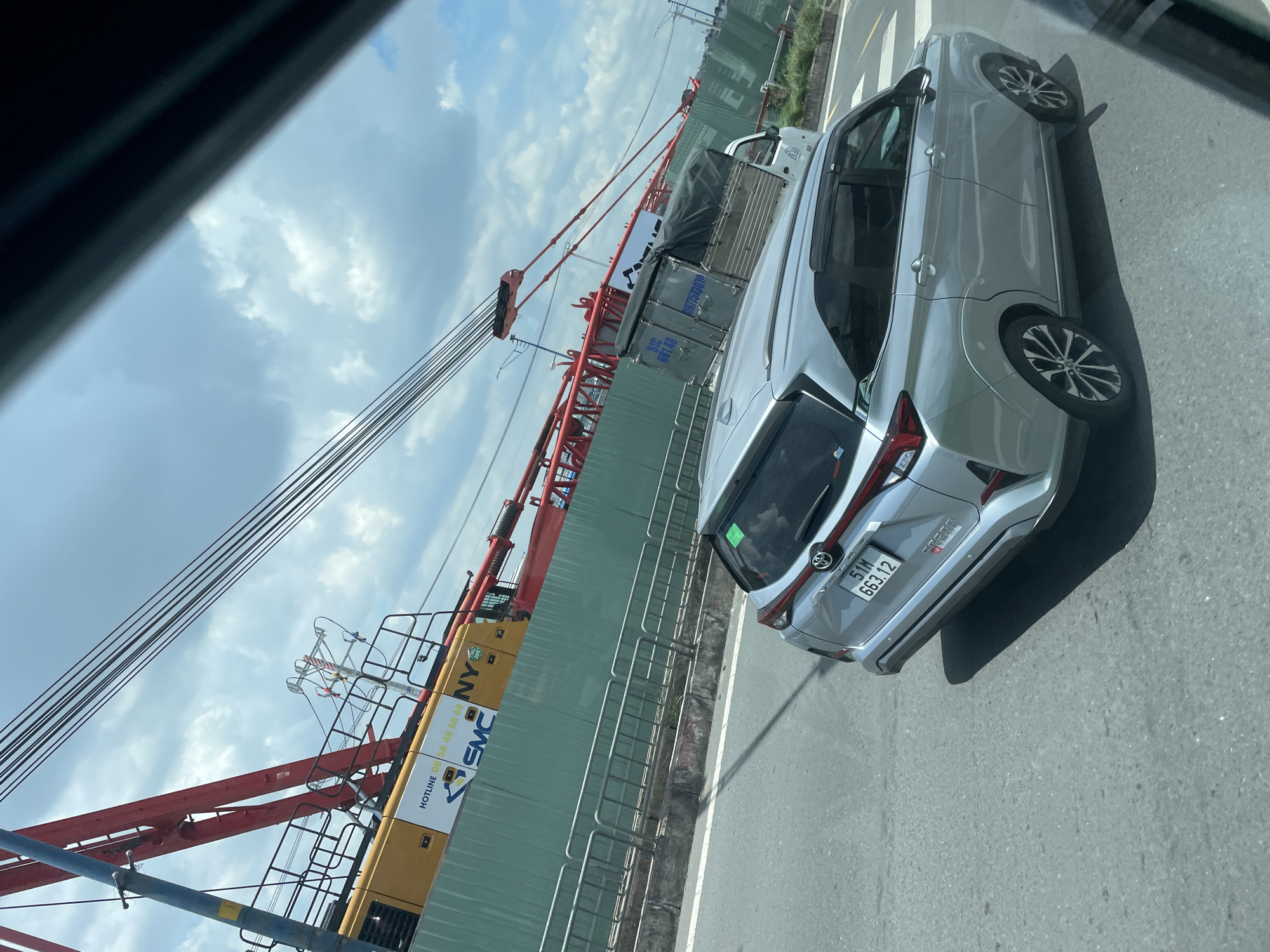 A silver sedan is captured from a rear-quarter angle, driving on an asphalt road in what appears to be Thuận An, Vietnam. The car's license plate reads "51M 663.12". In the background, a large red and yellow construction crane is visible, positioned behind a green fence. The sky is partly cloudy. The scene suggests a typical urban environment with ongoing construction.
