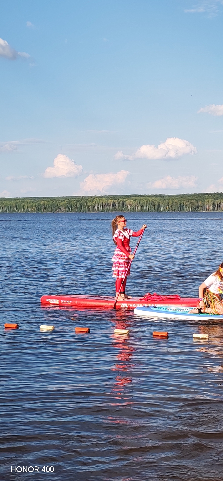 A young girl in a red and white costume stands on a red paddleboard, holding a paddle. She is looking to her right with a slight smile. Another person, partially visible on a blue paddleboard to the right, is also present. The paddleboards are on a body of water, likely a lake or river, with a line of buoys in the foreground. In the background, a dense line of green trees forms the horizon, beneath a blue sky with scattered white clouds. The scene appears to be set on a sunny day, possibly in the late afternoon, given the lighting and reflections on the water. The overall mood is peaceful and recreational. The text "HONOR 400" is visible in the bottom left corner. The location context is Chaikovsky, Russia.
