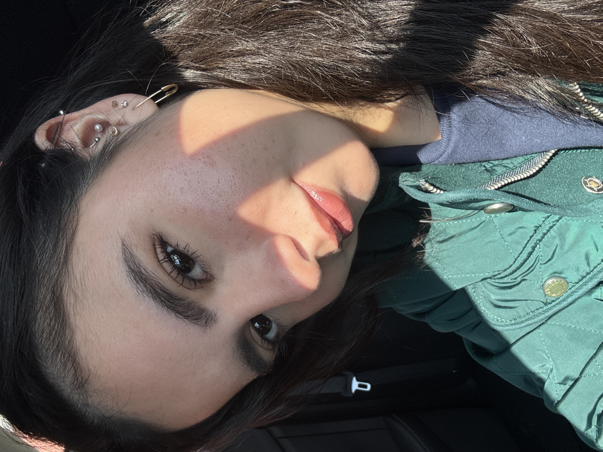 A close-up, rotated image shows a young woman with dark hair and brown eyes. She is wearing a dark green quilted jacket over a navy blue top. The woman has multiple ear piercings in her left ear, including a safety pin acting as an earring. Sunlight is casting a shadow across her face. The location and city are not discernible from the image.