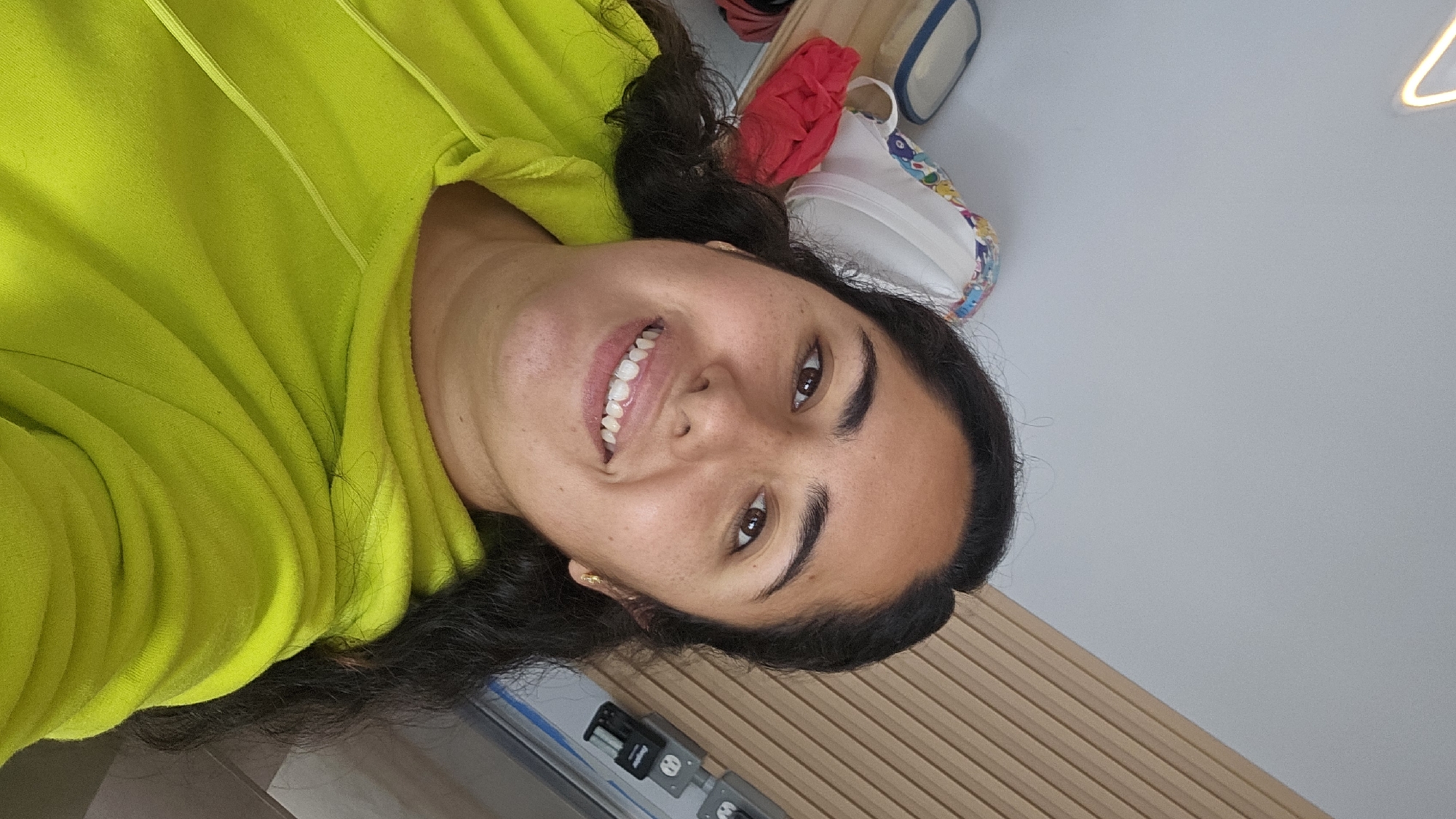 A young woman with dark hair and brown eyes, wearing a yellow hoodie, smiles at the camera. Her head is positioned at the center of the image, slightly tilted. Behind her, a wall with light brown vertical stripes is visible. To the upper left, there is a glimpse of what seems to be a shelf with a red item, a white item with colorful designs, and a light blue item. The background is a neutral light grey. The image does not provide any information that would indicate the location in Puebla, Mexico.