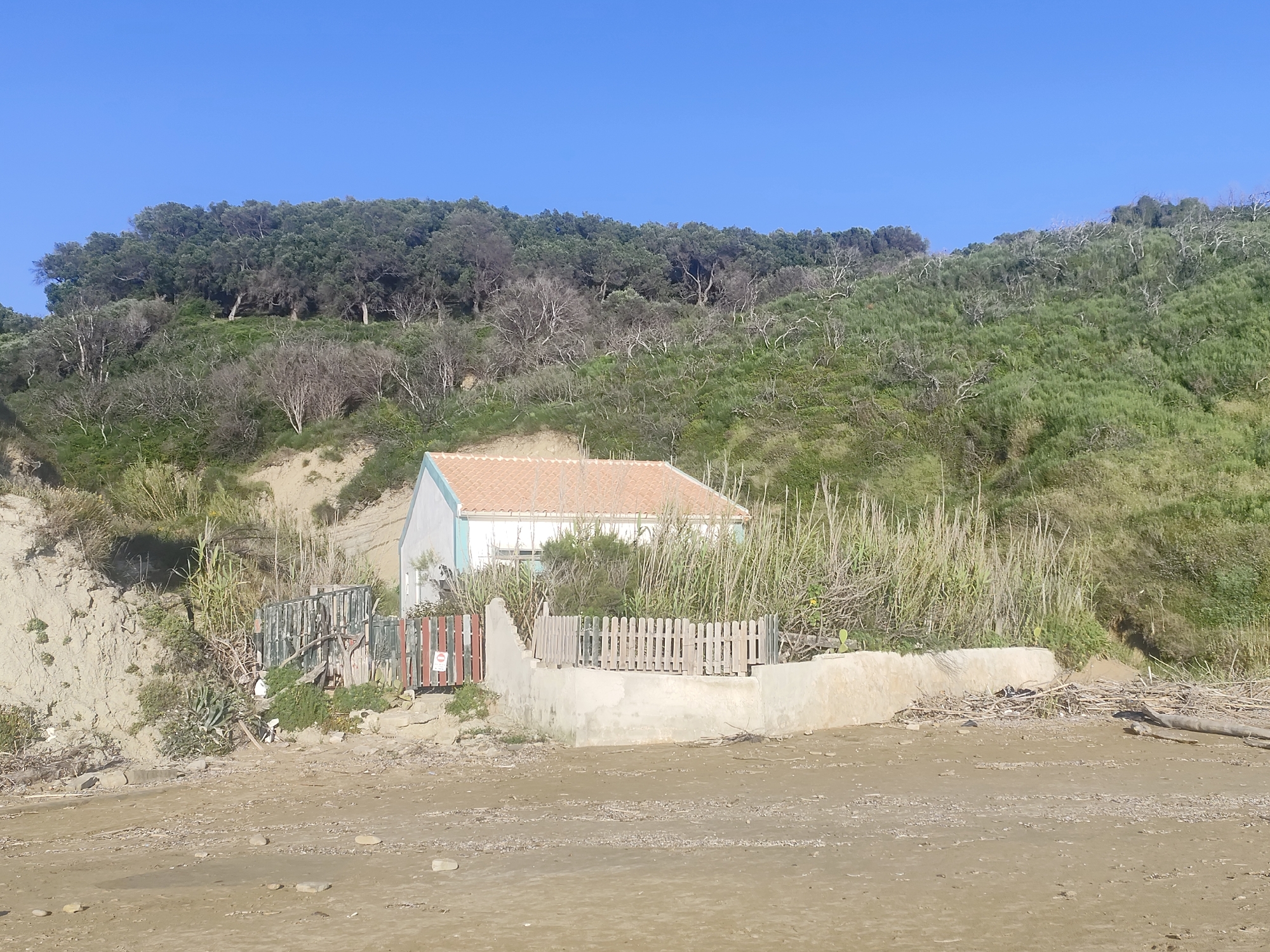 The image depicts a small, single-story house situated on a beach, nestled against a hillside. The house has a light-colored exterior with a reddish-brown tiled roof. A low concrete wall and a wooden fence surround the house. The hillside behind the house is covered with green vegetation and some trees. The beach in front of the house is sandy and appears to be wet, with some debris scattered on it. The sky above is clear and blue. The location is Unknown, Unknown.