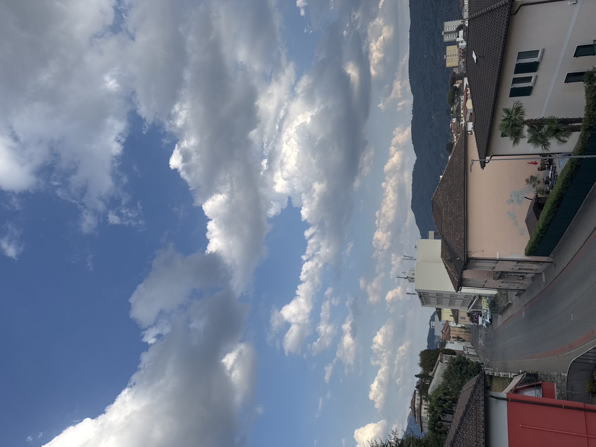 The image shows a view of a street in Balerna, Switzerland, with buildings and a backdrop of mountains. The sky is partly cloudy with a mix of blue sky and white and gray clouds. In the foreground, there are buildings with tiled roofs, one of which is painted red. A paved street runs through the scene, with some cars parked along the side. There are trees and greenery visible. In the mid-ground, several multi-story buildings are visible, including a tall, light-colored building with antennas on top. In the background, rolling, tree-covered hills or mountains form the horizon. The overall impression is of a residential or commercial area within a town nestled in a hilly or mountainous region.