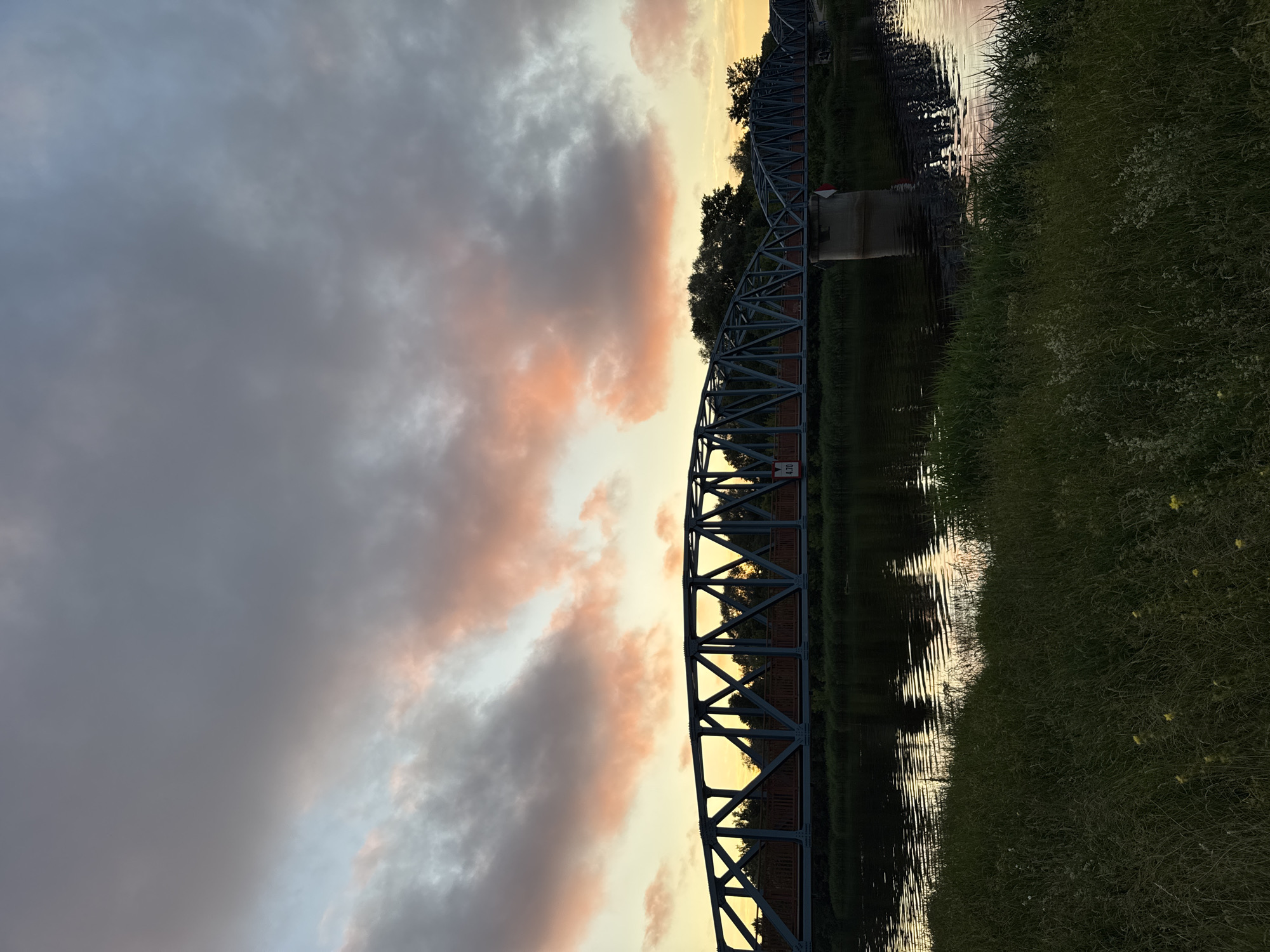 The image shows a steel truss bridge spanning across a body of water, likely a river, in Carnikava, Latvia. The sky is filled with dramatic clouds tinged with pink and orange, suggesting either sunrise or sunset. The water reflects the colors of the sky and the bridge. Lush green grass and some wildflowers line the foreground on the riverbank. Trees are visible on the far bank of the river behind the bridge. There are no people or animals visible in the image. A sign with "4,70" is visible on the bridge.