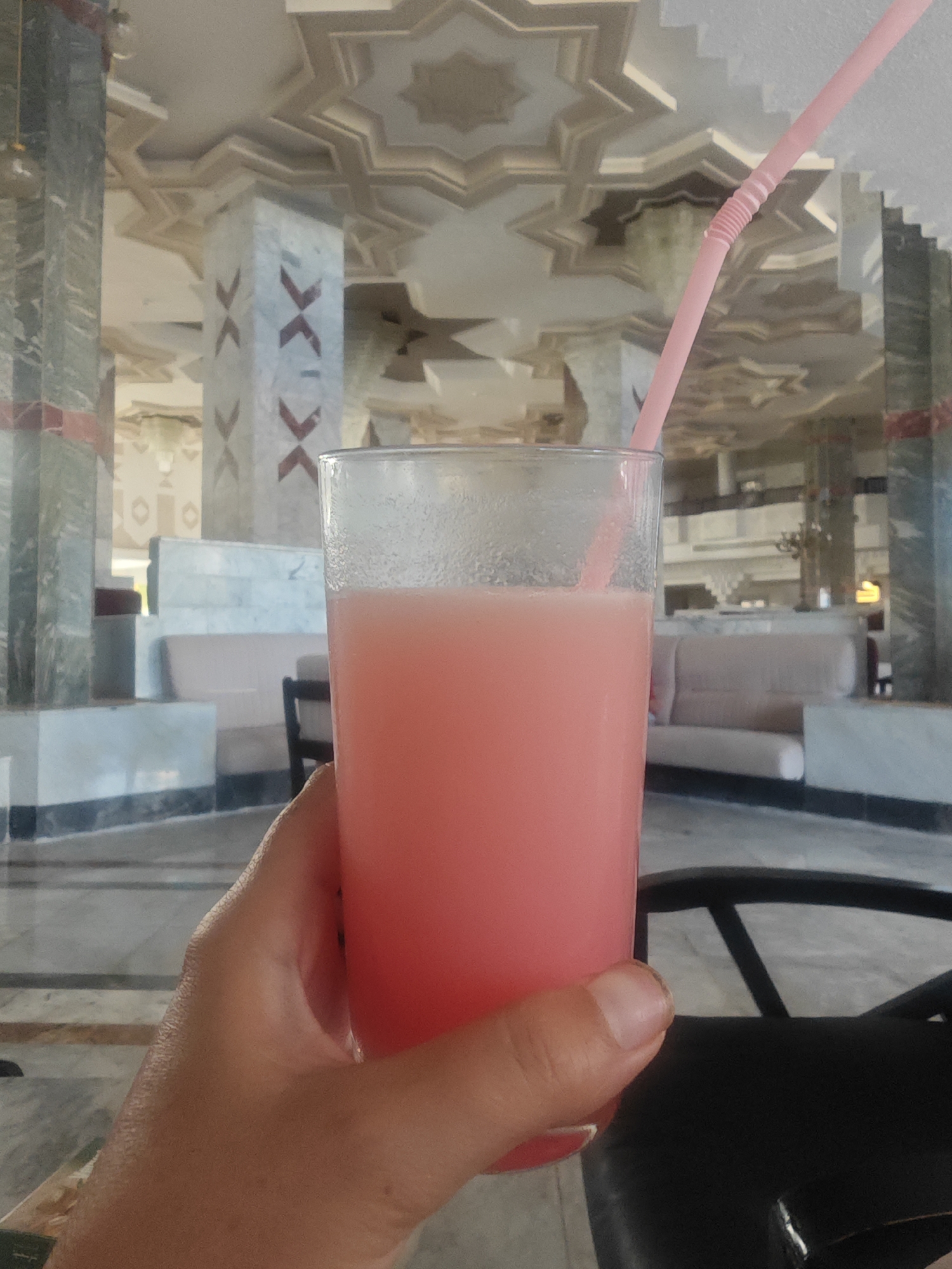 A close-up shot focuses on a hand holding a glass of pink beverage with a pink straw. The background reveals a lavishly decorated interior, possibly a hotel lobby or a lounge area, characterized by ornate ceilings with star-shaped patterns, marble-clad columns with intricate designs, and plush seating. The overall aesthetic suggests a luxurious and possibly resort-like environment. The location is identified as Hiboun, Tunisia.