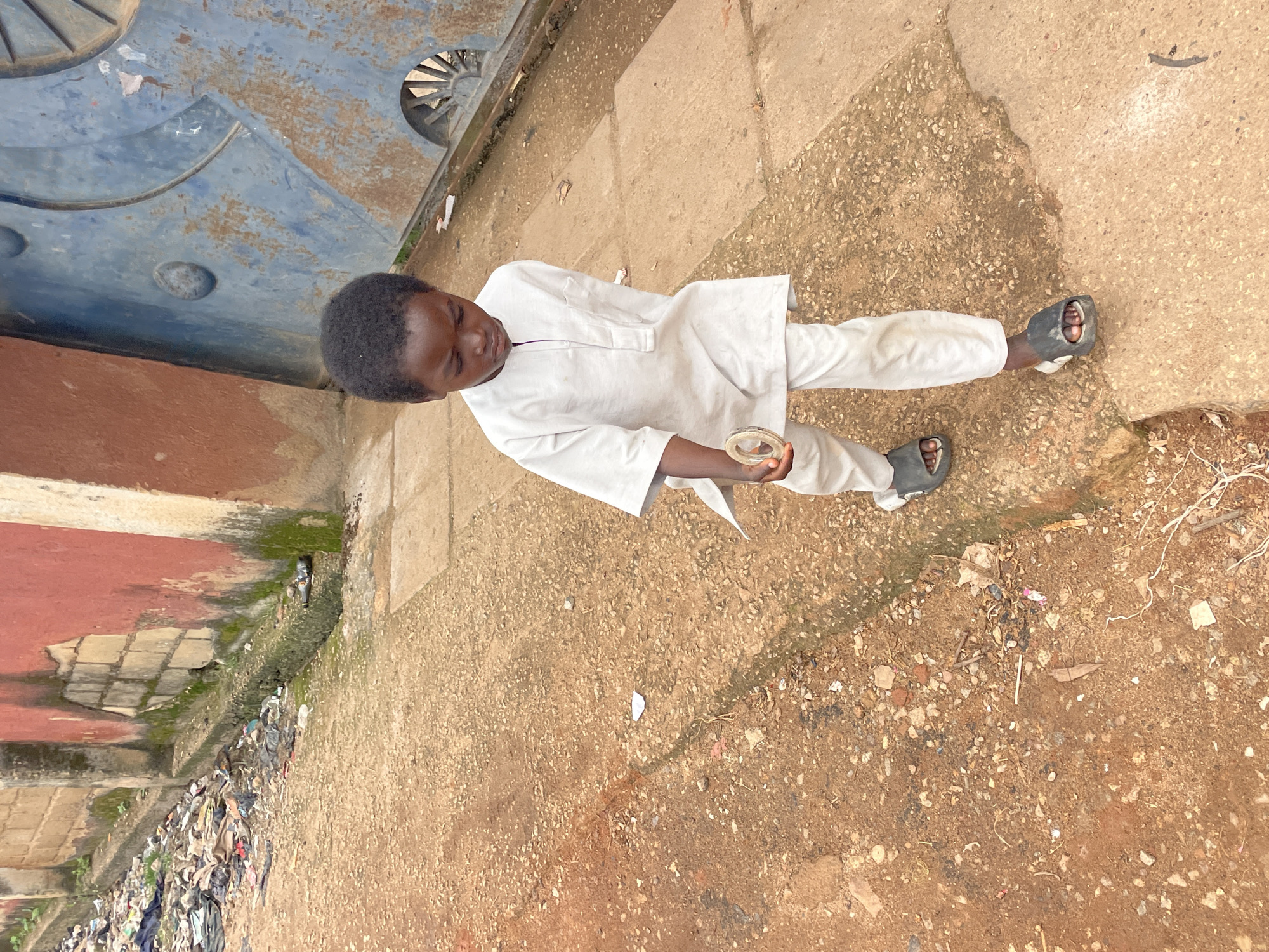 A young boy walks on a dirt path in front of a red and cream colored building. He is wearing a white tunic and pants. He is holding a small, circular object in his right hand. To the left of the boy, there is a pile of trash. To the right of the boy, there is a blue metal gate. The scene appears to be in Jos, Nigeria.