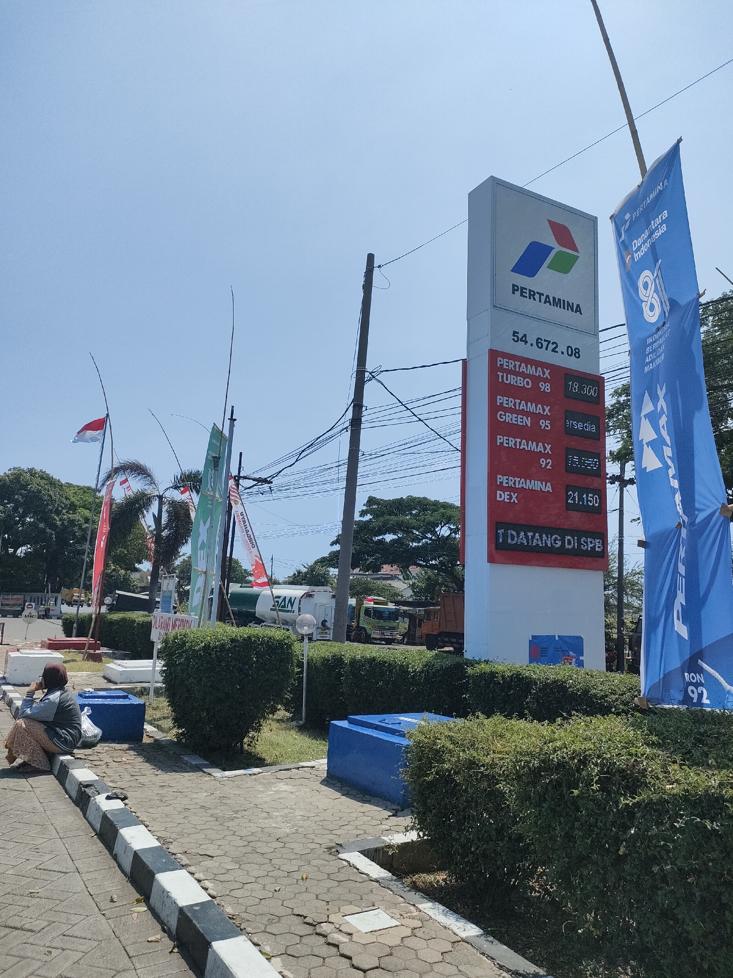 The image depicts a Pertamina gas station in Curahdringu, Indonesia. A tall, white sign displays the Pertamina logo and fuel prices. The prices listed are Pertamax Turbo 98 at 18,300, Pertamax Green 95 available, Pertamax 92 at 13,750, and Pertamina Dex at 21,150. Below the prices, a red sign reads "T DATANG DI SPB". Two blue flags with Pertamina branding are present, one on the left and a larger one on the right. In the foreground, a woman sits on a sidewalk, facing away from the camera, with a bag beside her. A black and white striped curb runs along the sidewalk. Green hedges line the front of the gas station, and several tall poles with flags are visible, including an Indonesian flag. In the background, a white Pertamina tanker truck is parked, and other vehicles are present, suggesting an active gas station environment. The sky is clear and blue, indicating a sunny day.