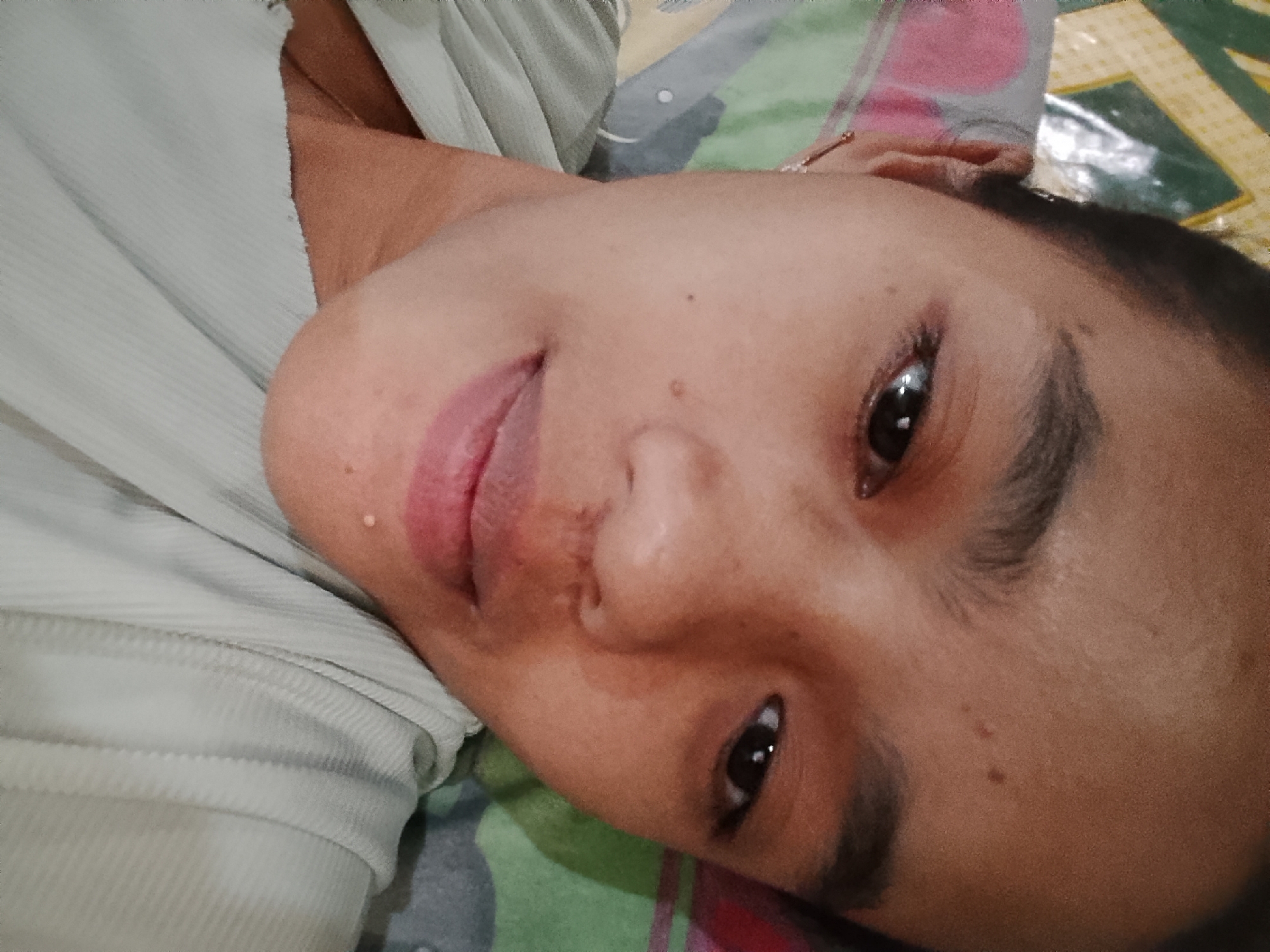An upward-facing, close-up, portrait-style photograph captures a young Indonesian person lying on their back. The subject's head is tilted, and their face is angled towards the camera. They are wearing a light-colored, ribbed garment. The background consists of a patterned fabric, likely a bedspread or mat, featuring green, pink, and yellow geometric designs. A small, clear droplet is visible on the subject's upper lip, possibly sweat or a reflection. The lighting is soft and diffuse, indicating an indoor setting. The image provides a detailed view of the subject's facial features, including their eyes, nose, and mouth, as well as a portion of their shoulder and garment. The context suggests a personal photograph taken in a private space within Besuki, Indonesia.