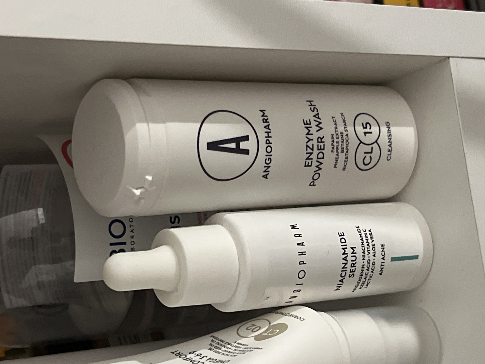 This is a close-up, eye-level, indoor photograph of skincare products on a shelf. The background is out of focus but appears to be a cabinet or closet with other items. The main subjects are two white cylindrical bottles from the brand ANGIOPHARM.
The bottle on the left is a dropper bottle, labeled "NIACINAMIDE SERUM" and "ANTI ACNE." Its label also lists ingredients: "ANGIOGENIN • NIACINAMIDE • AZELAIC ACID • VITAMIN C • LACTIC ACID • ALOE VERA." The dropper cap is white.
The bottle on the right is taller and appears to be a powder wash. It is labeled "ANGIOPHARM ENZYME POWDER WASH." Beneath this, it lists ingredients: "PAPAIN PINEAPPLE EXTRACT BETAINE RICE&TAPIOCA STARCH." There is also a circular logo with "CL 15" inside, and the word "CLEANSING" below it. The cap is white and opaque.
A third, partially visible white tube is to the left of the ANGIOPHARM bottles. Its label has text including "COMFORT," "OMEGA 3-6-9," and "CORNEOTHE..."
The image does not contain any information that would allow for the identification of its location in Kazan, Russia. The scene depicts a collection of personal care items, likely in a bathroom or storage area.