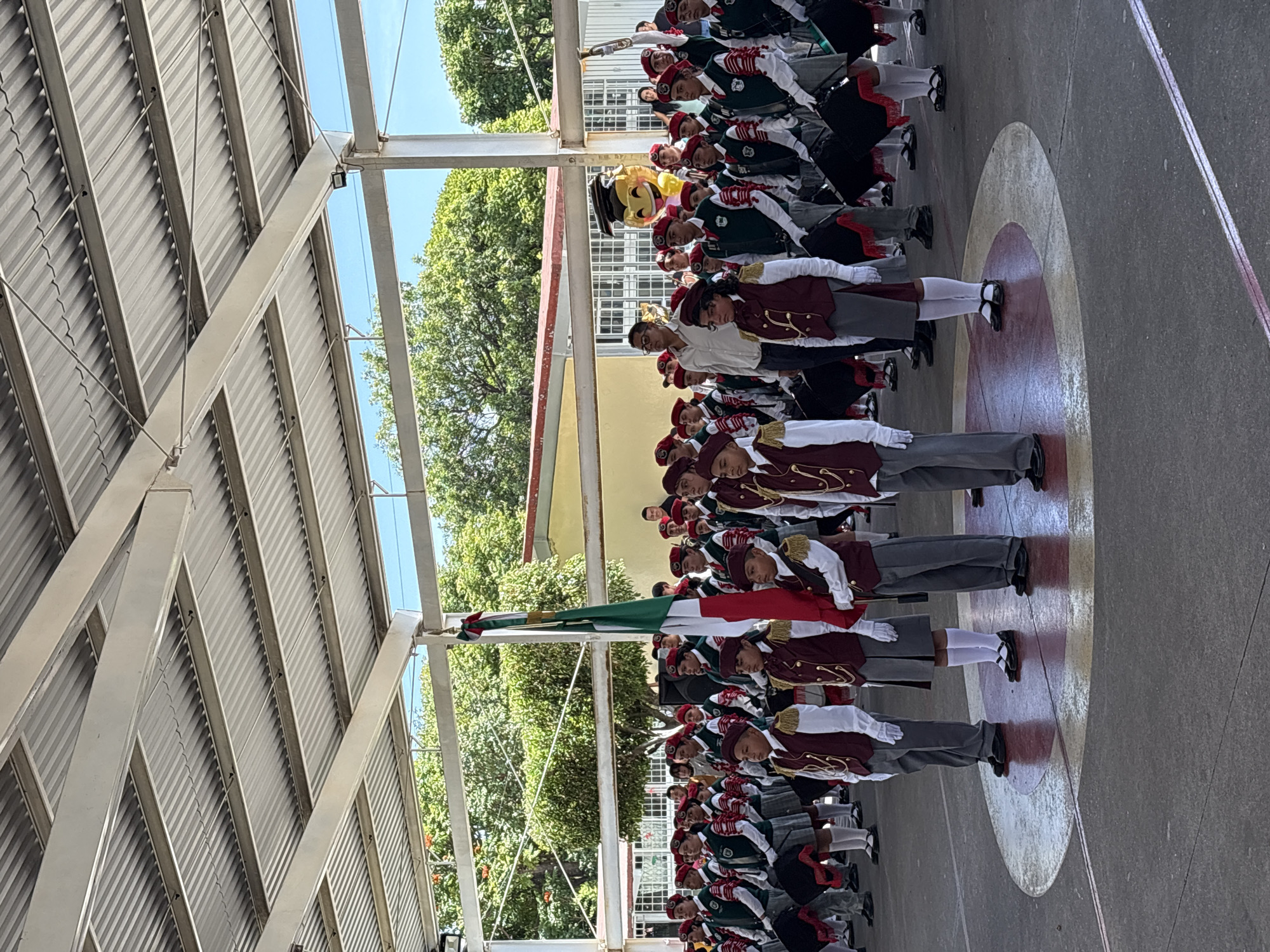 In Tejalpa, Mexico, a group of students, likely a marching band, stand in formation on a paved outdoor area. They are dressed in coordinated uniforms: maroon vests with gold trim over white shirts, gray skirts or pants, and red berets. Many are holding drums or drumsticks, and some are wearing white gloves. A Mexican flag is prominently displayed and held by one of the students. In the background, trees and a building are visible, and the scene is covered by a large, white corrugated metal roof supported by metal beams. A yellow balloon with a graduation cap and a smiling face is visible in the upper right portion of the image. The students appear to be engaged in a formal ceremony or parade, standing at attention.