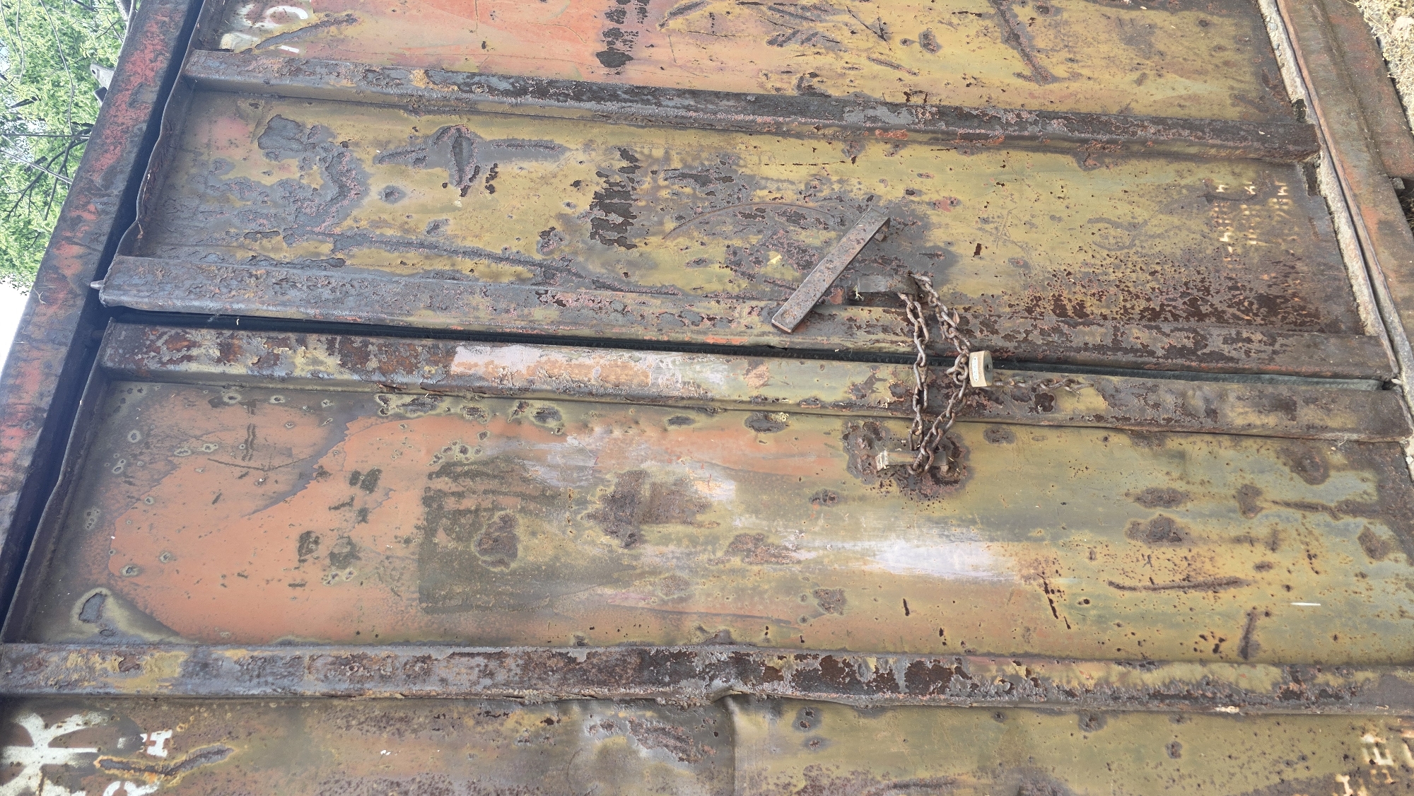 The image shows a close-up of a heavily rusted metal door or gate. A chain and padlock secure the door. Patches of original paint, in shades of orange and yellow, are visible beneath the rust. The door is constructed with horizontal metal bars reinforcing the flat panels. The image provides no information that can be used to determine location in Blagoevgrad, Bulgaria.
