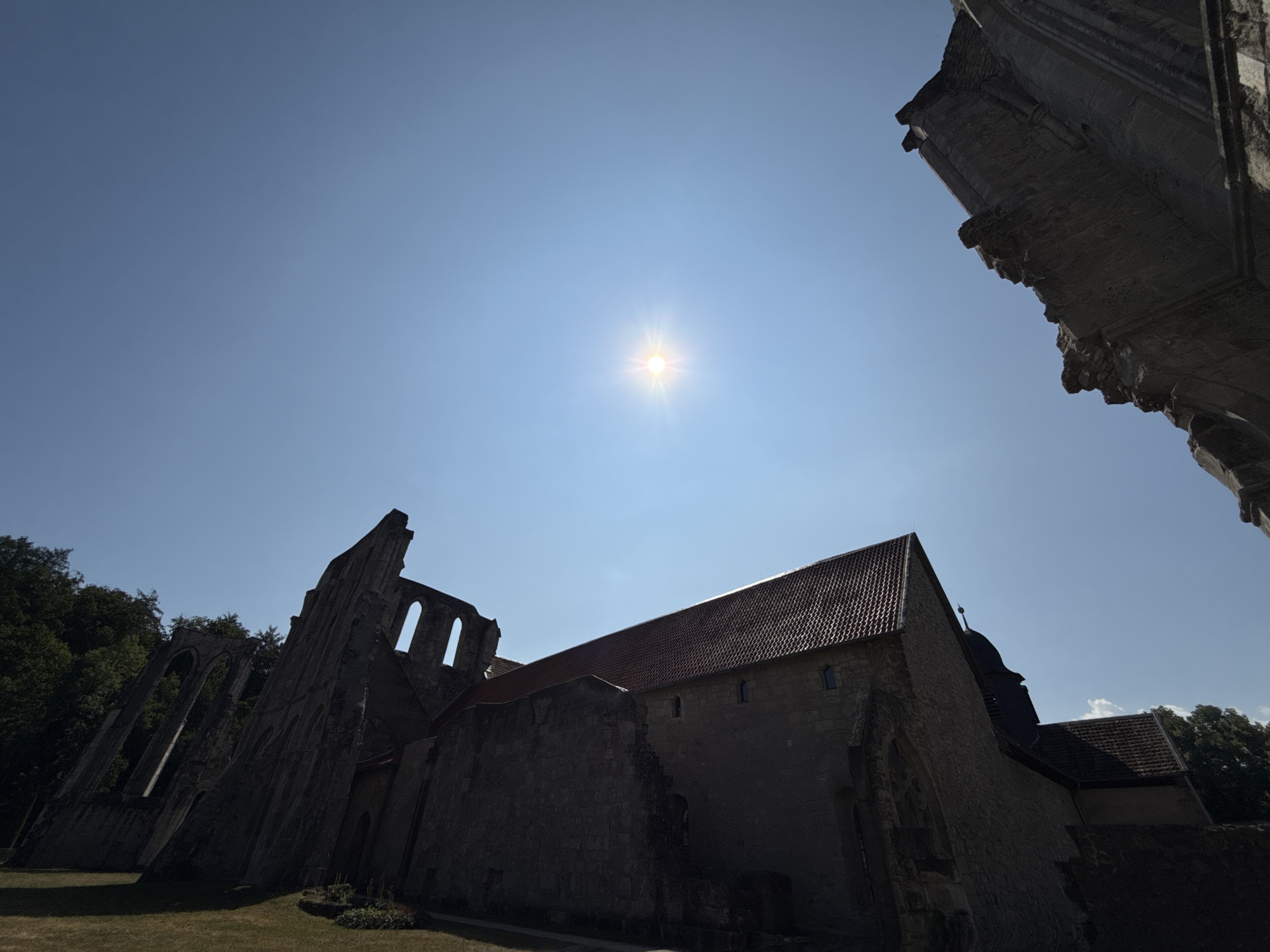 The image depicts the ruins of the Walkenried Abbey in Walkenried, Germany, against a clear blue sky. The sun is visible, casting a bright glare. The stone ruins are silhouetted against the sky, with some sections appearing more intact than others. One part of the abbey has a red-tiled roof. Trees are visible in the background on the left and right sides. The ground in front of the ruins is covered in grass. The perspective is low, looking upwards at the architecture.