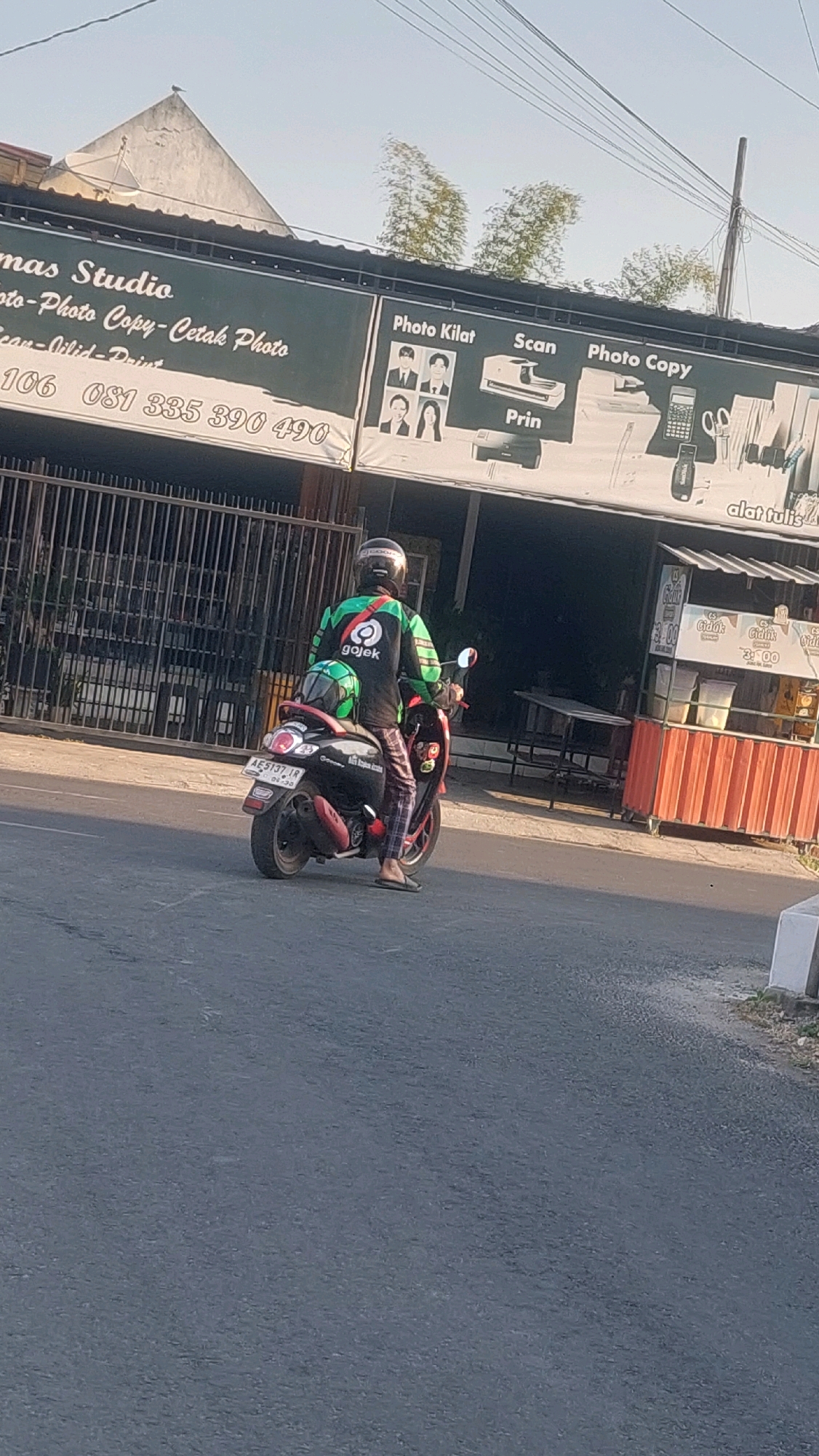 In Ronowijayan, Indonesia, a Gojek rider on a scooter travels down a road in front of a building with signage for a photo studio and copy services. The rider, wearing a helmet and the distinctive green and black Gojek uniform, is seated on a black scooter with a green helmet secured to the back. The building's facade features a dark green banner displaying services like "Photo Kilat," "Scan," "Photo Copy," and "Prin." The street is paved, and a metal fence runs along the side of the building, partially obscuring a seated area with tables and chairs. In the background, a white building with a triangular roof and a satellite dish is visible. The sky is clear and bright, suggesting daytime.