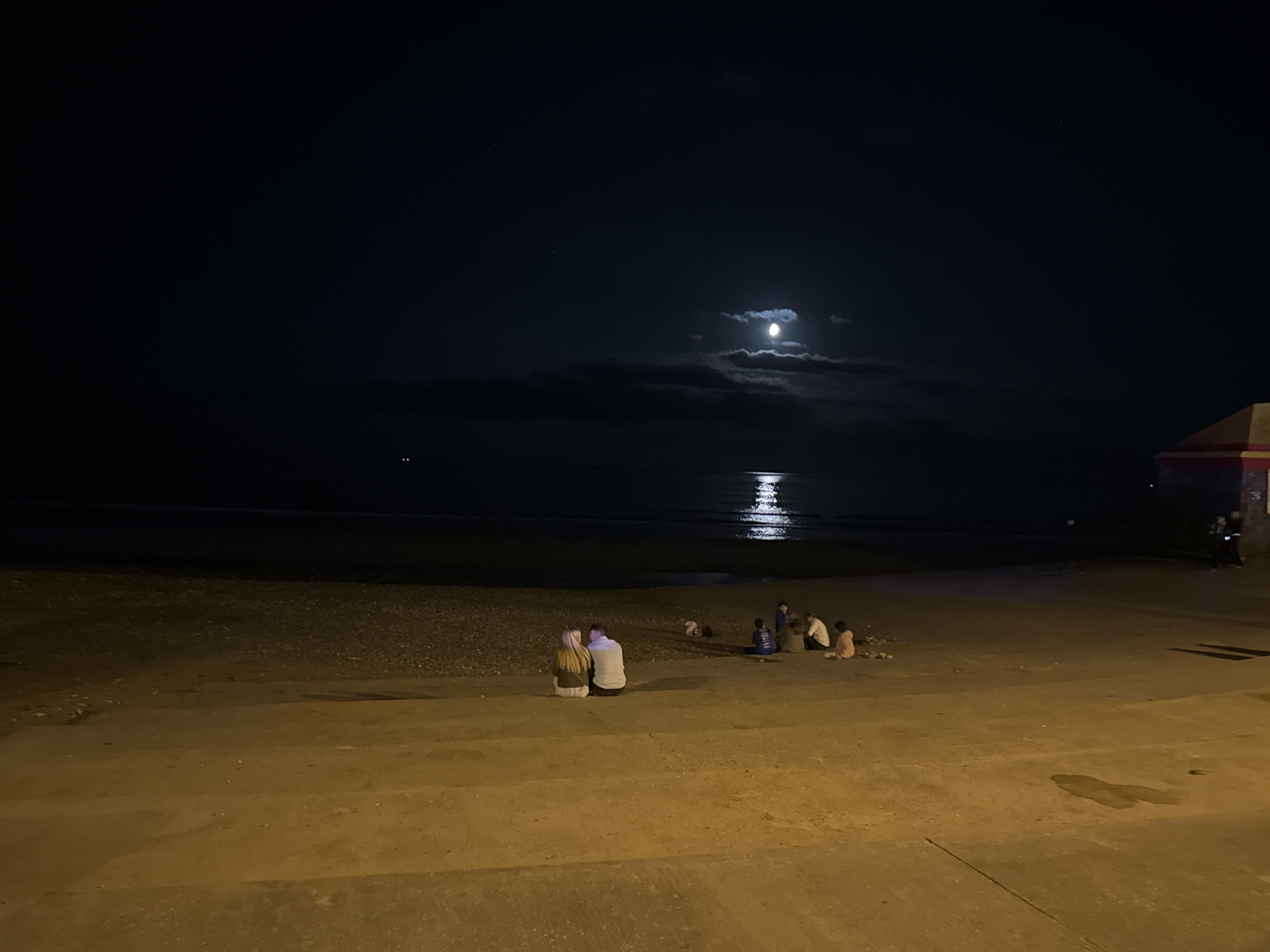 At night, a wide, dark expanse of sea meets a pebble beach. The moon, partially obscured by clouds, casts a bright reflection on the water. In the foreground, a paved walkway borders the beach. Several people are gathered on the beach and walkway. On the left, a couple sits close together on the walkway, facing the sea. Further back on the beach, a group of individuals, including children, are seated or crouching. On the far right, a portion of a building with red and yellow accents is visible, possibly a structure at the amusement pier in Tramore, Ireland. The scene is illuminated by ambient light from the moon and possibly artificial lighting from the pier area, casting long shadows.