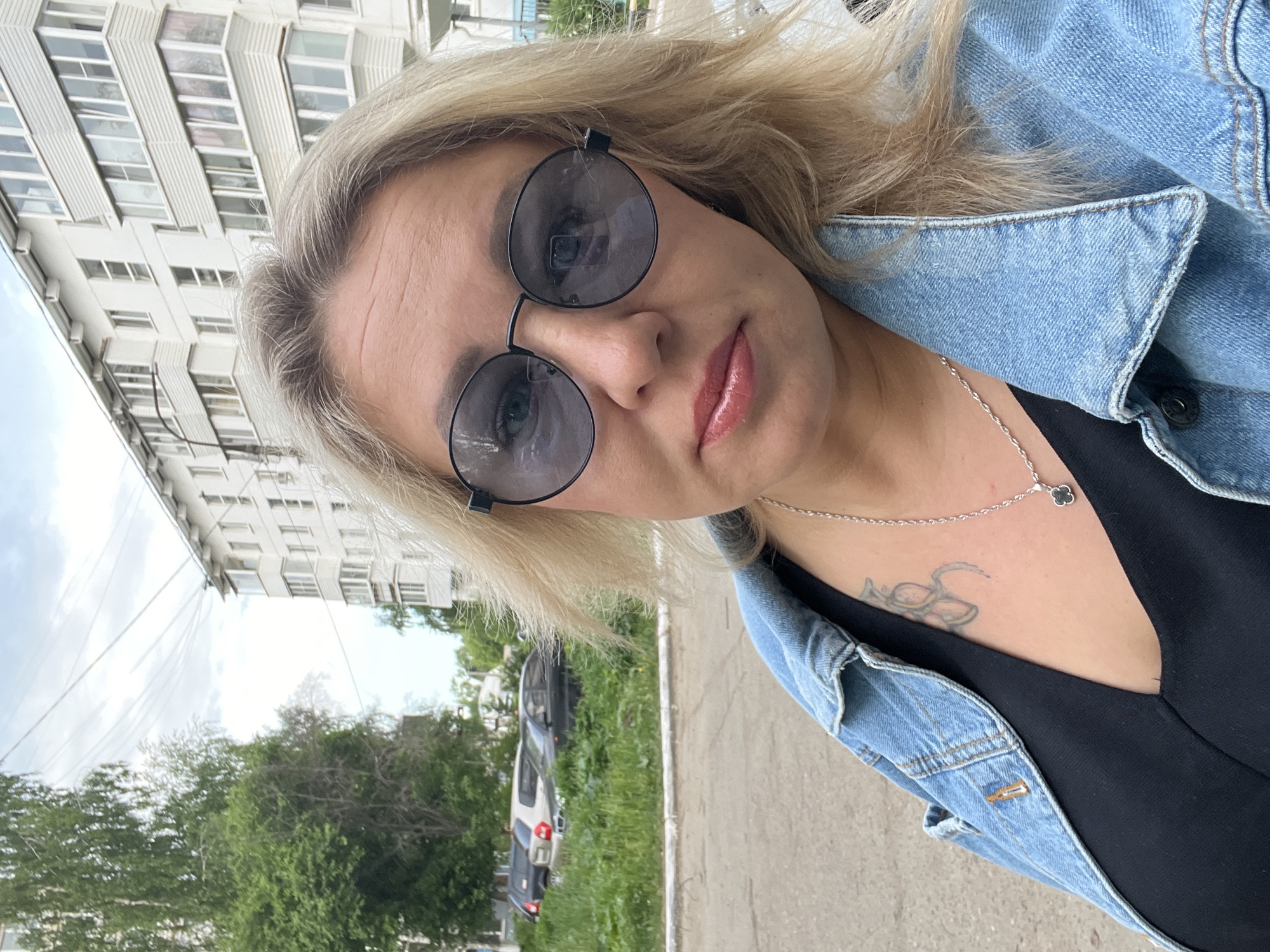 A fair-skinned blonde woman wearing sunglasses and a denim jacket takes a selfie. She is also wearing a black top and a silver necklace with a clover pendant. A tattoo is visible on her upper chest. The background shows a white apartment building, trees, parked cars, and a paved sidewalk. The location appears to be Chaikovsky, Russia.