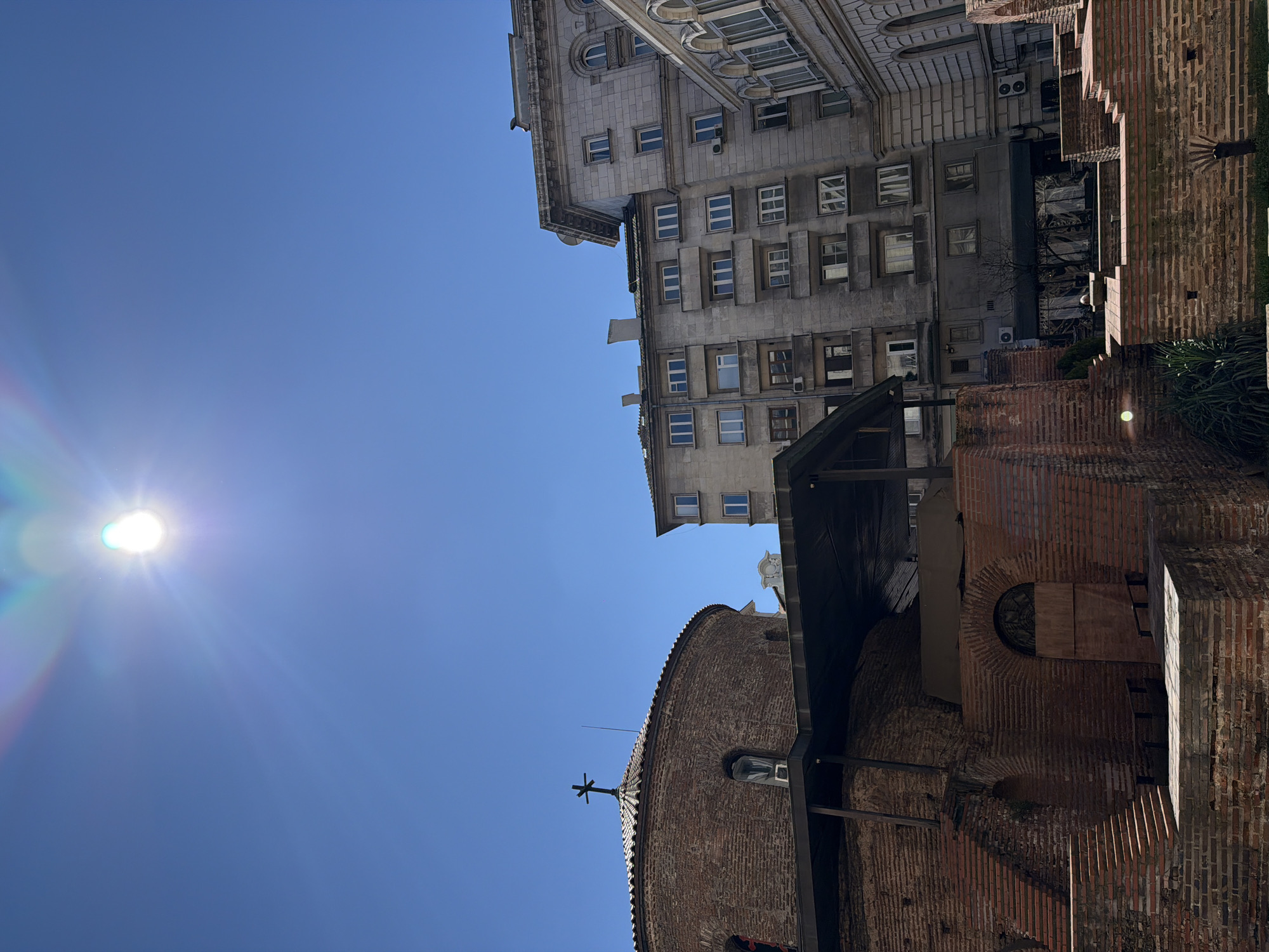 The image shows the St. George Rotunda church in Sofia, Bulgaria. The church is a small, red brick structure with a dome topped by a cross. The building is surrounded by other buildings, including a large, multi-story stone building with many windows. The sky is blue and clear, with the sun visible in the upper left corner.
