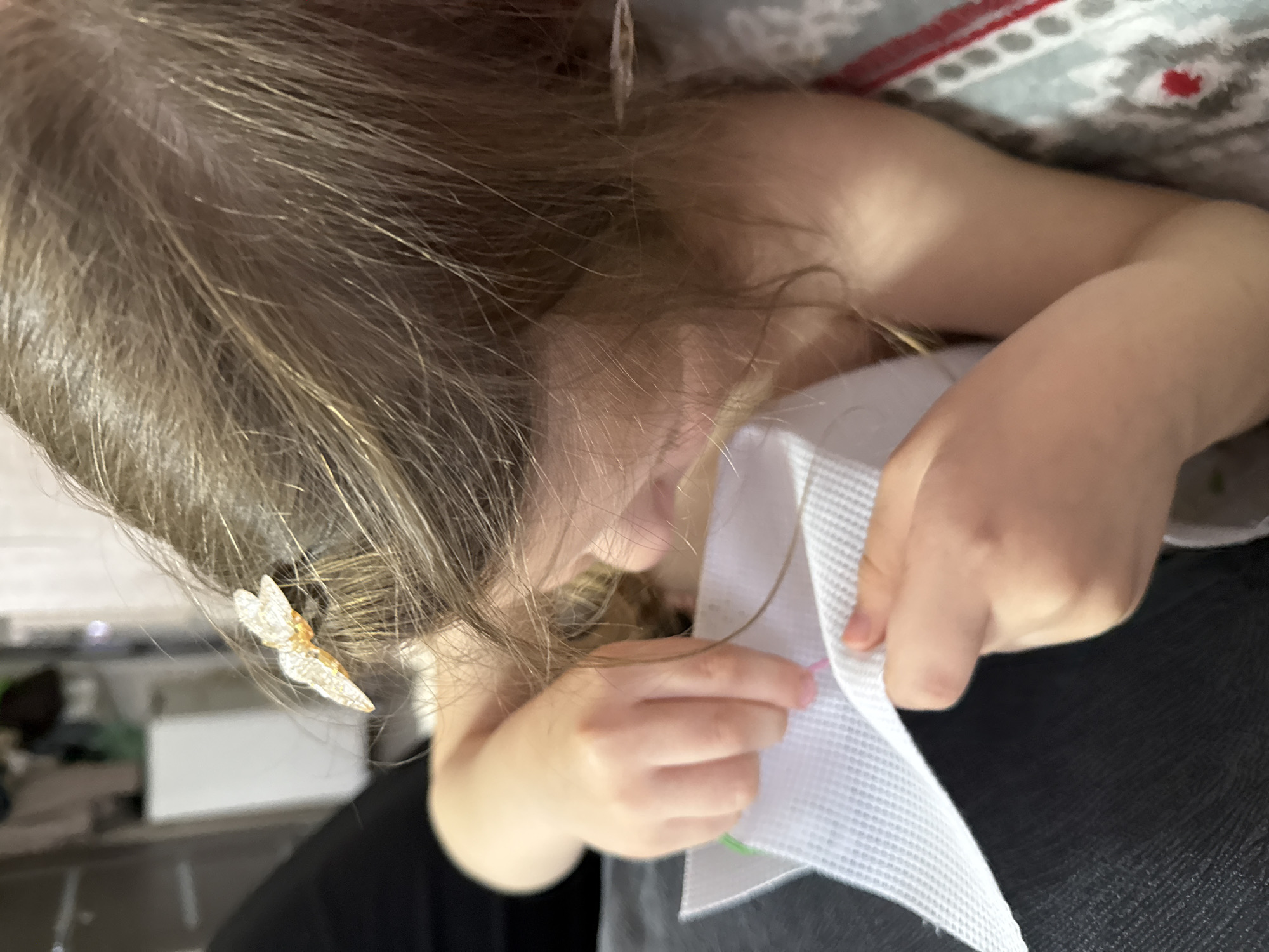 A child is crouched and engrossed in working with a piece of white plastic canvas, threaded with pink and green embroidery floss. The child has brown hair with blonde highlights and is wearing a butterfly-shaped hair clip. The child is working on a black surface, possibly seated in a chair. A patterned blanket is visible in the background. The location is not discernible and cannot be confirmed as Кипчак, Russia.
