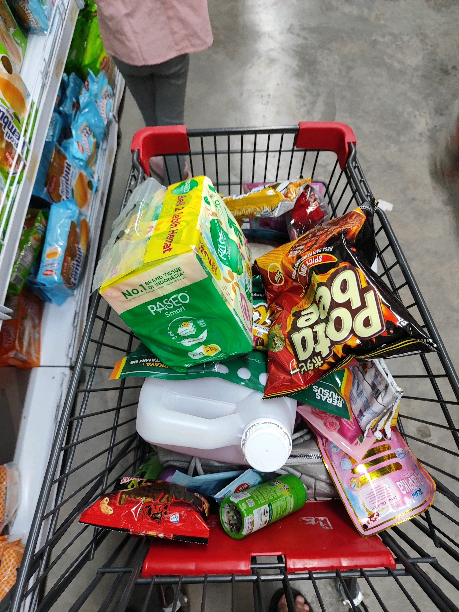 A first-person perspective shot captures a shopping cart filled with groceries in what appears to be a supermarket aisle in Sukowiryo, Indonesia. The cart is laden with various packaged goods, including a prominent yellow and green box of Paseo Smart tissue, several bags of chips, a white plastic container, and a red plastic item in the bottom. The background shows shelves stocked with more products, suggesting a retail environment. A person's legs, wearing gray pants and a pink top, are visible at the top of the frame, indicating they are pushing the cart. The floor is a concrete surface.