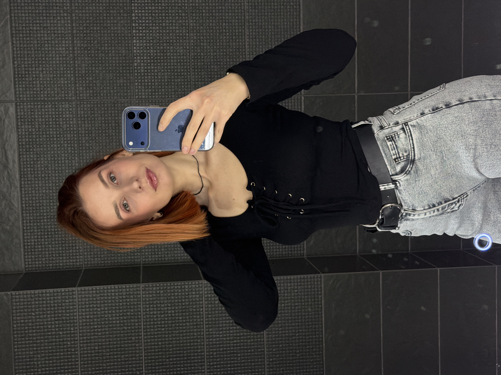 A woman with red hair is taking a selfie in what appears to be a bathroom. She is holding a blue iPhone in her right hand. She is wearing a black long-sleeved top with corset lacing at the neckline and light-wash jeans with a black belt. The bathroom has a tiled floor and tiled walls in various shades of gray. The location is not discernible from the image.