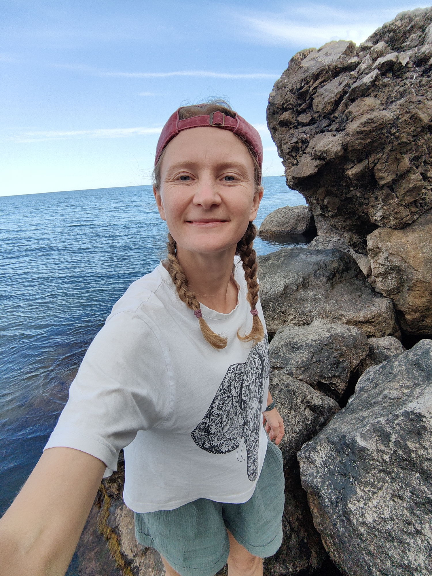 A woman with blonde hair in two braids, wearing a red baseball cap, a white t-shirt with an elephant print, and green shorts, smiles at the camera. She is standing on rocks next to the sea. The scene appears to be in Utios, Ukraine, characterized by a rocky coastline and clear blue water under a partly cloudy sky.