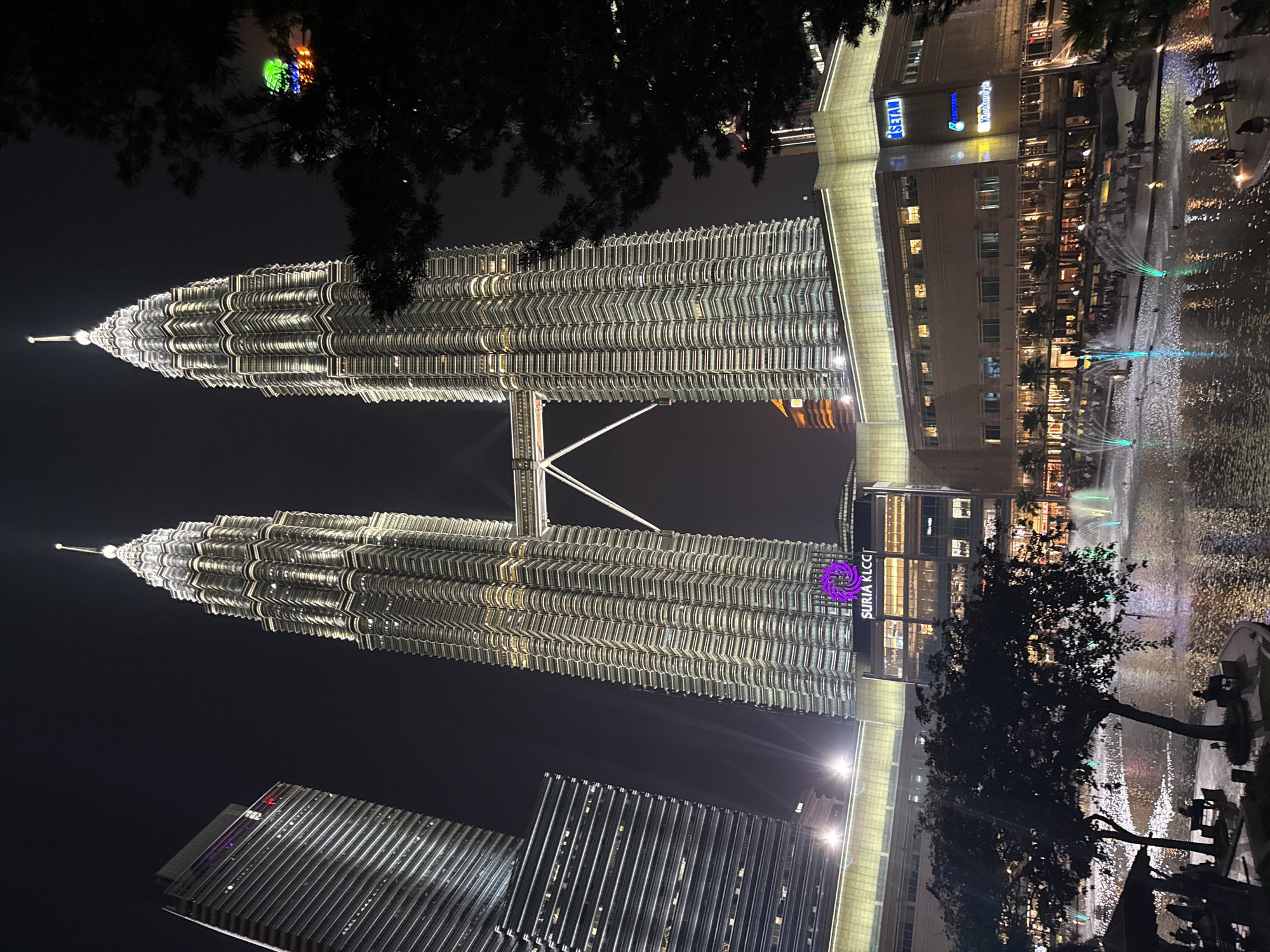 The image captures a nighttime view of the iconic Petronas Twin Towers in Kuala Lumpur, Malaysia, rotated 90 degrees clockwise. The two towering skyscrapers, brightly illuminated, dominate the frame, showcasing their intricate, segmented metallic facades and pointed spires against a dark night sky. A skybridge connects the two towers at a middle height.
In the foreground, the dark silhouettes of tree branches obscure parts of the upper sections of the towers, adding depth to the composition. To the right of the towers, a large water feature or fountain is visible, with multiple jets of water illuminated by vibrant blue and green lights, creating a dynamic display. Adjacent to the fountain is a multi-story building, likely a commercial center, with many windows lit up, revealing interiors.
Visible text on this building includes "iSETAL" in white, "Kopitiam" in yellow, and another logo with a 'K' inside a circle. On the base of one of the Petronas Towers or an adjacent building, a prominent purple logo and text read "SURIA KLCC," indicating the shopping mall associated with the towers. Another modern high-rise building with reflective surfaces is partially visible in the lower left, also illuminated. The clear night sky and the abundance of artificial light highlight the urban grandeur of the scene. No discernible people are visible in the image.