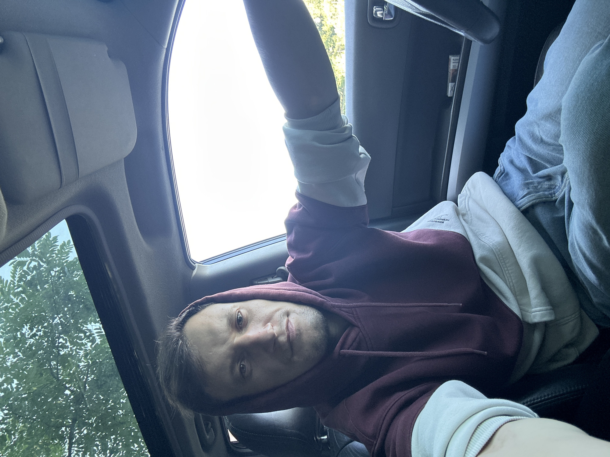 A man with fair skin and short, dark brown hair is in the driver's seat of a car. He is wearing a maroon hooded sweatshirt with cream-colored sleeves and blue jeans. His left arm is resting on the steering wheel. The car has a sunroof, through which green tree leaves are visible. The interior of the car is dark, with a light-colored dashboard and door panels. The man is looking directly at the camera with a neutral expression. The scene appears to be captured during the daytime, with bright light coming from outside. The location is Makiv, Ukraine.