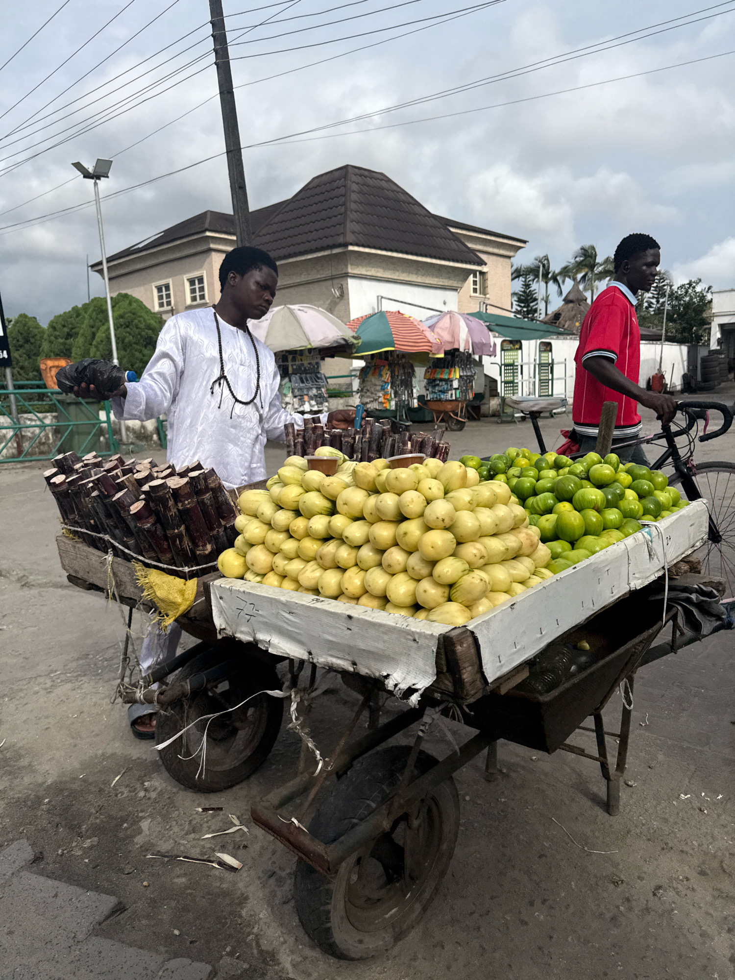 The image depicts a street scene in Ado, Nigeria. Two young men are associated with a fruit and sugar cane cart. The man on the left, dressed in a white garment and black beaded necklace, holds a dark plastic bag in his left hand and appears to be interacting with the cart's contents. The cart itself is laden with produce, including large piles of light yellow fruits and bundles of dark sugar cane stalks. To the right of the cart, a second man, wearing a red t-shirt, stands near a bicycle and is partially obscured by the cart. Behind the cart, several market stalls with colorful umbrellas are visible, suggesting a marketplace setting. Buildings with tiled roofs and palm trees are in the background, under a cloudy sky. The ground is paved with concrete and appears somewhat worn.